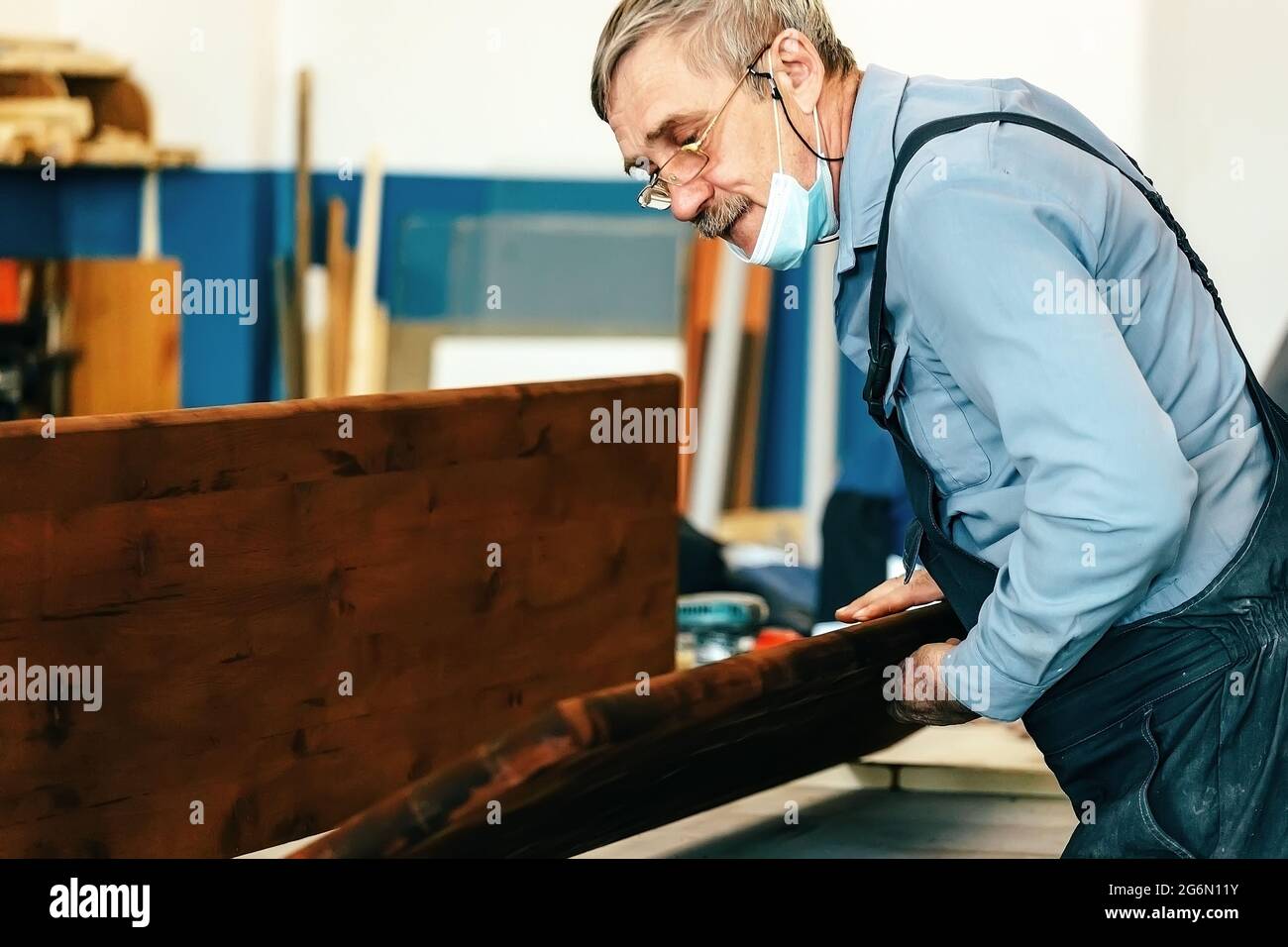 Ein grauhaariger Zimmermann mit Brille arbeitet mit Holz auf einer Werkbank  in einer Schreinerei. Ein Rentner in einer medizinischen Maske bei der  Arbeit. Beschäftigung und Arbeit ein älterer Mann in Arbeitskleidung  Stockfotografie -