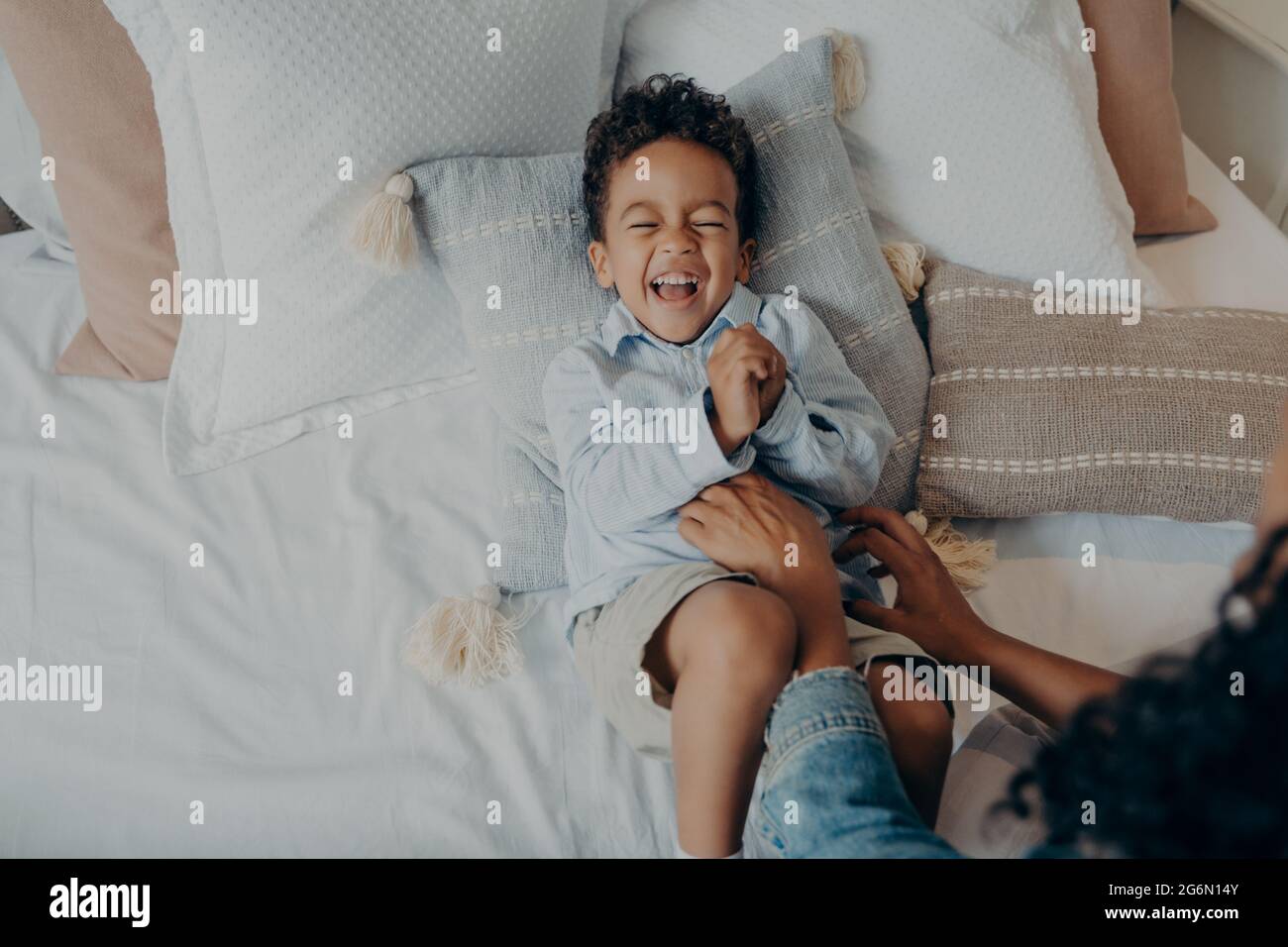 Entzückender kleiner Sohn, der auf Kissen im Bett lag und lachte, während seine Mutter ihn kitzelte Stockfoto