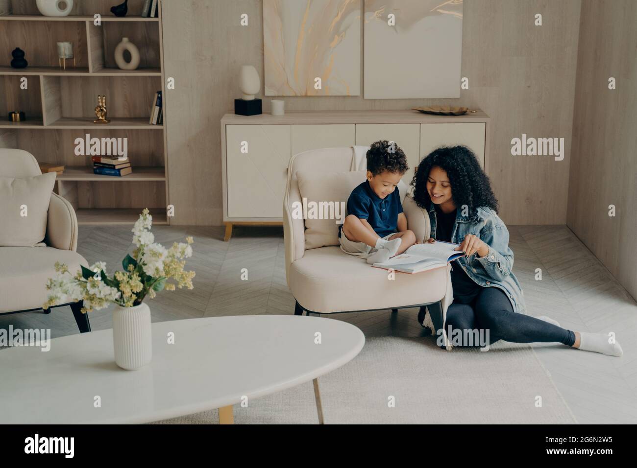 Kleiner gemischter Rennjunge, der mit seiner liebevollen Mutter liest, während er zu Hause im Wohnzimmer Zeit zusammen genießt Stockfoto