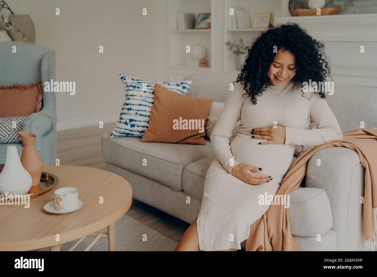 Lächelnd schöne schwanger Mischling Frau auf der Couch sitzen und berühren ihren Bauch Stockfoto