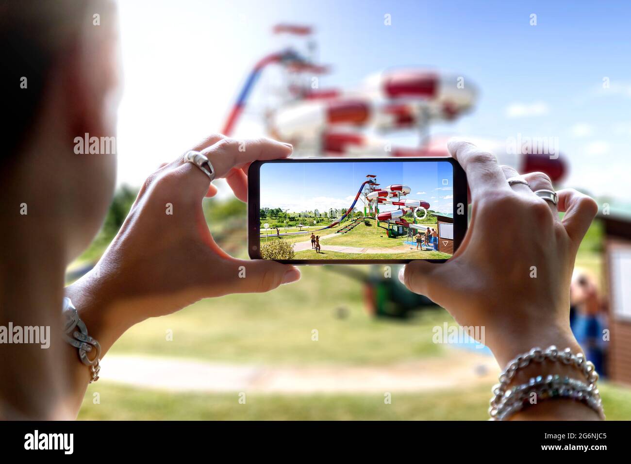 Frau in einem Wasserpark, die mit ihrem Mobiltelefon ein Foto gemacht hat. Fotografieren einer großen Wasserrutsche mit der Kamera eines Smartphones. Stockfoto