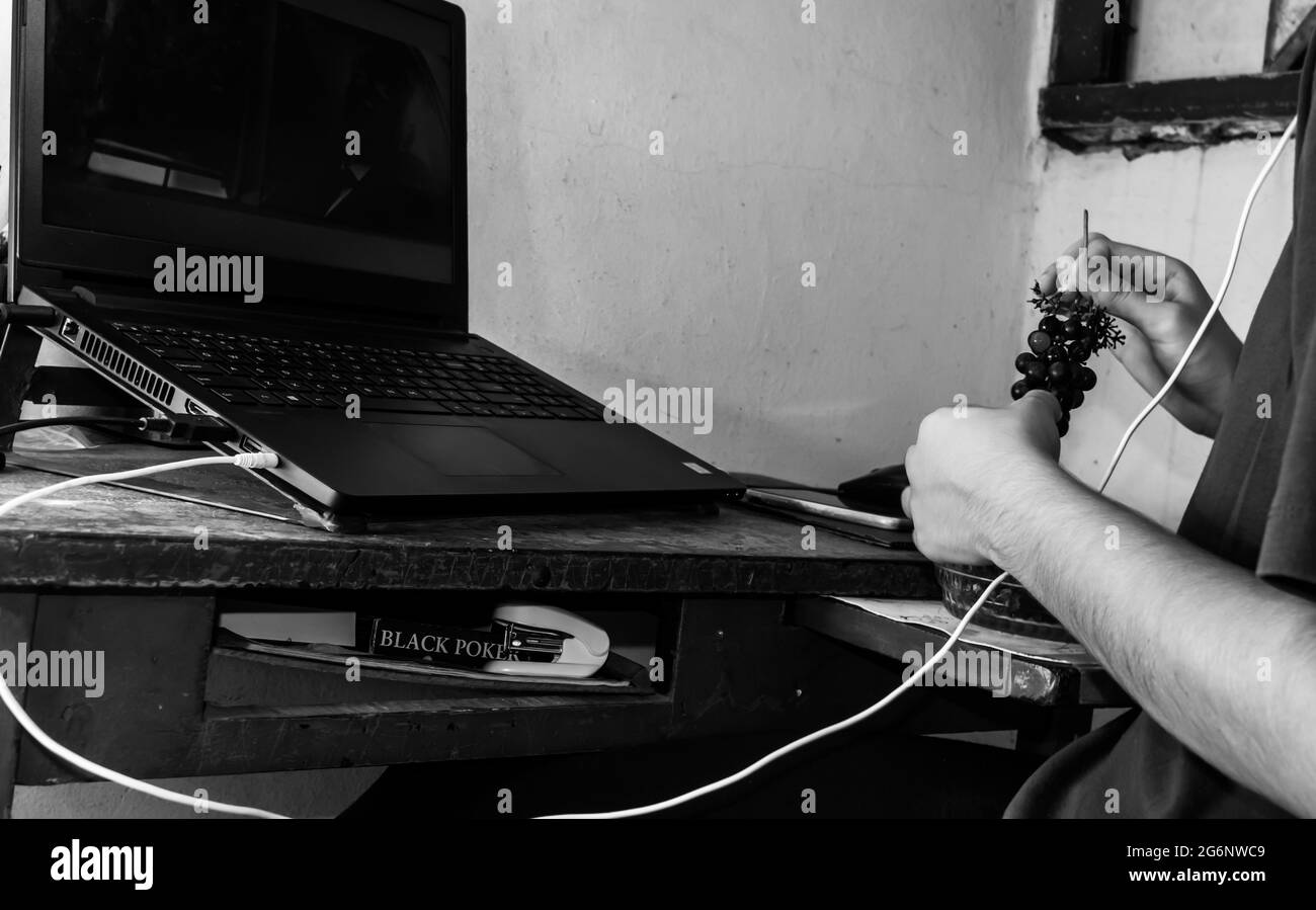 Ein alter Schreibtisch mit einem schwarzen Laptop darauf, darunter eine Schachtel mit schwarzen Steckkarten, und vor dem Laptop sitzt jemand, der Grapefrui trägt Stockfoto