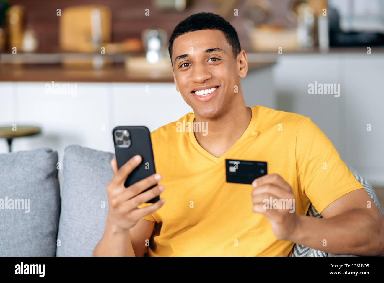 Positiv attraktiver hispanischer Kerl, in orangefarbenem T-Shirt, verwendet sein Smartphone und seine Kreditkarte, um für Online-Einkäufe zu bezahlen, Lieferung, eine Online-Transaktion zu tätigen, Zahlungen, lächelt glücklich. Online einkaufen Stockfoto