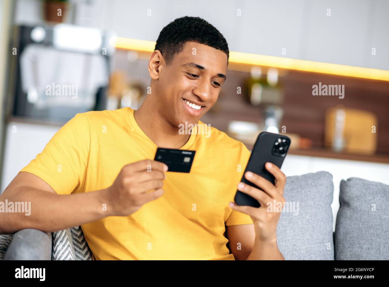 Fröhlicher, positiver hispanischer junger Mann, der im Wohnzimmer auf dem Sofa sitzt, in gelbem T-Shirt gekleidet, Smartphone und Kreditkarte zum Online-Shopping verwendet, Daten zur Zahlung eingibt, Online-Transaktionen vornimmt, lächelt Stockfoto