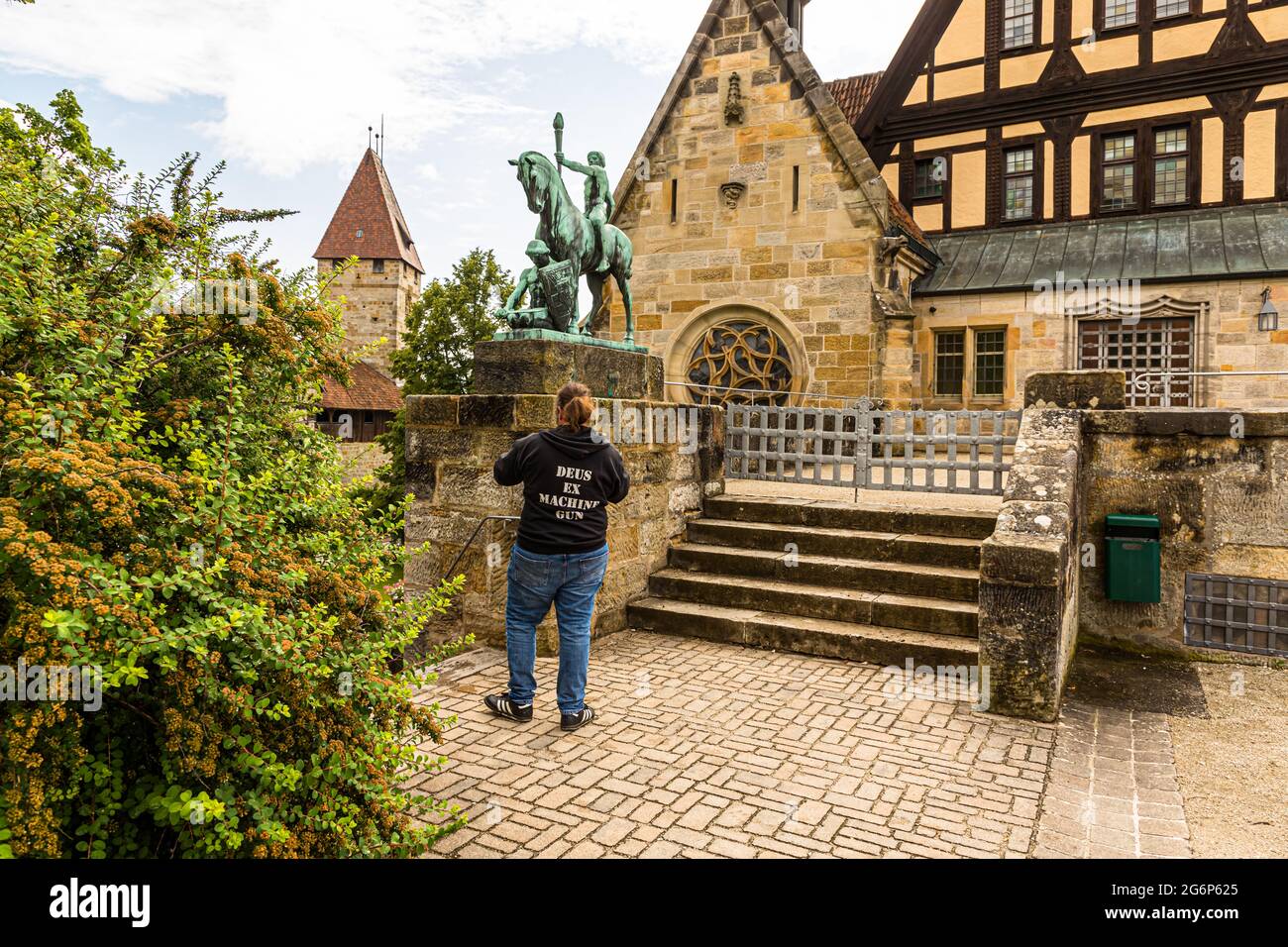 Das Reiterdenkmal "Licht und Kraft" in der Veste Coburg, Deutschland, erinnert an den monatelangen Aufenthalt Martin Luthers in der Festung im Jahr 1530. Der Bildhauer Hans Klett schuf die Bronzestatue 1913 zum 400. Jahrestag der Veröffentlichung der Thesen Luthers. Ein vor dem Denkmal stehender Besucher trägt ein T-Shirt mit der Aufschrift "Deus ex Machine Gun". Stockfoto