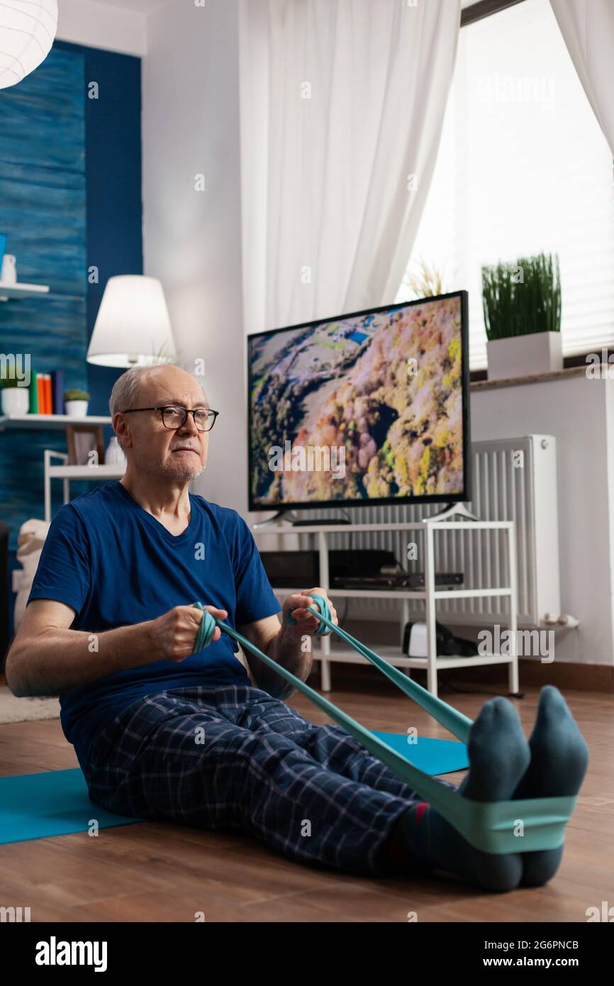 Seniorchef im Ruhestand sitzt auf einer Yogamatte und dehnt die Beine mit einem elastischen Band aus, um die Körperflexibilität zu trainieren. Rentner in Sportbekleidung abnehmen Gewicht beim Muskeltraining im Wohnzimmer Stockfoto
