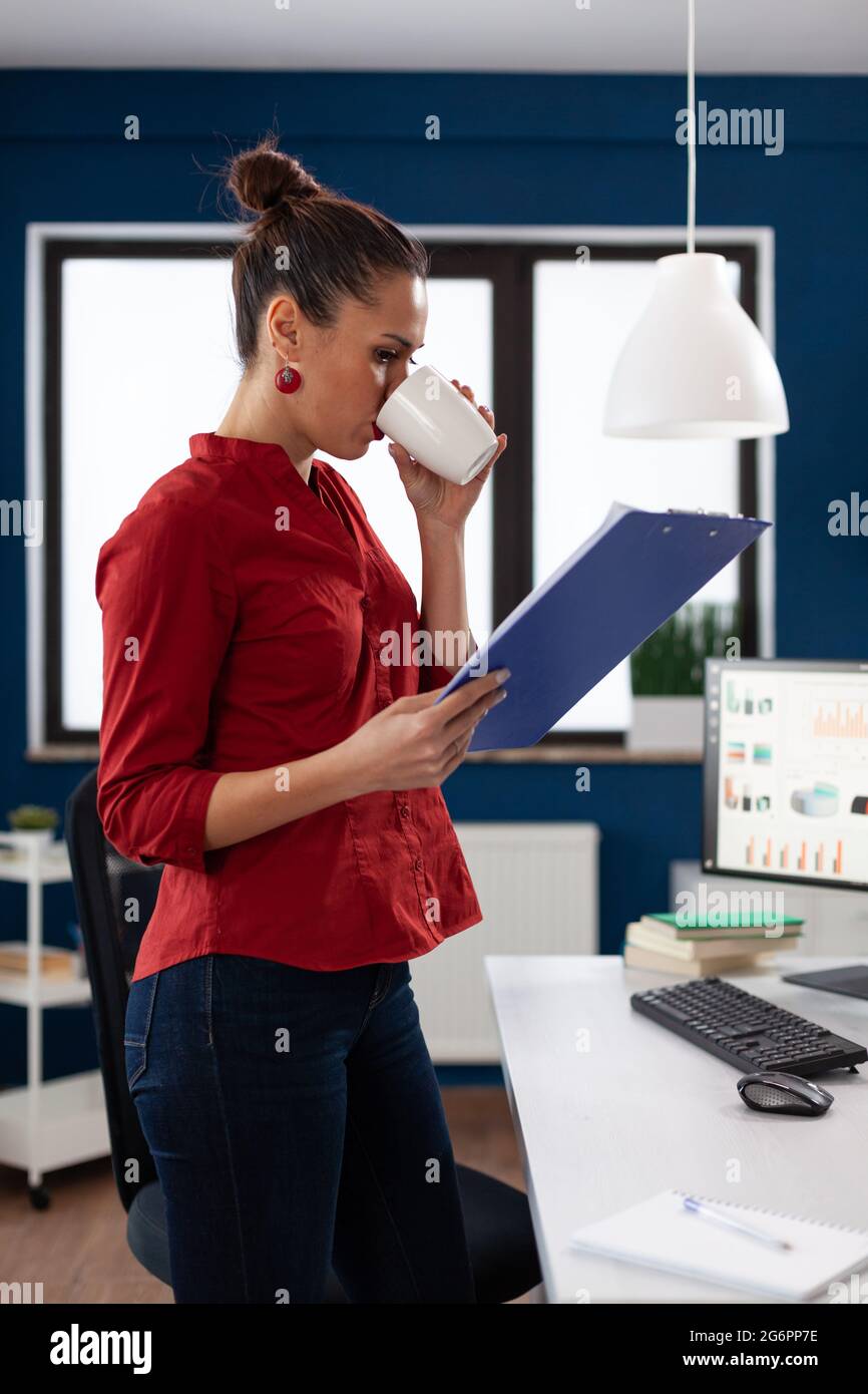 Erfolgreiche Geschäftsfrau im Büro des Unternehmens, die sich durch die Charts schaut und dabei eine Tasse leckeren Kaffee hält und jokiert. Gedankenvoll Unternehmer immer Doze von Koffein. Stockfoto