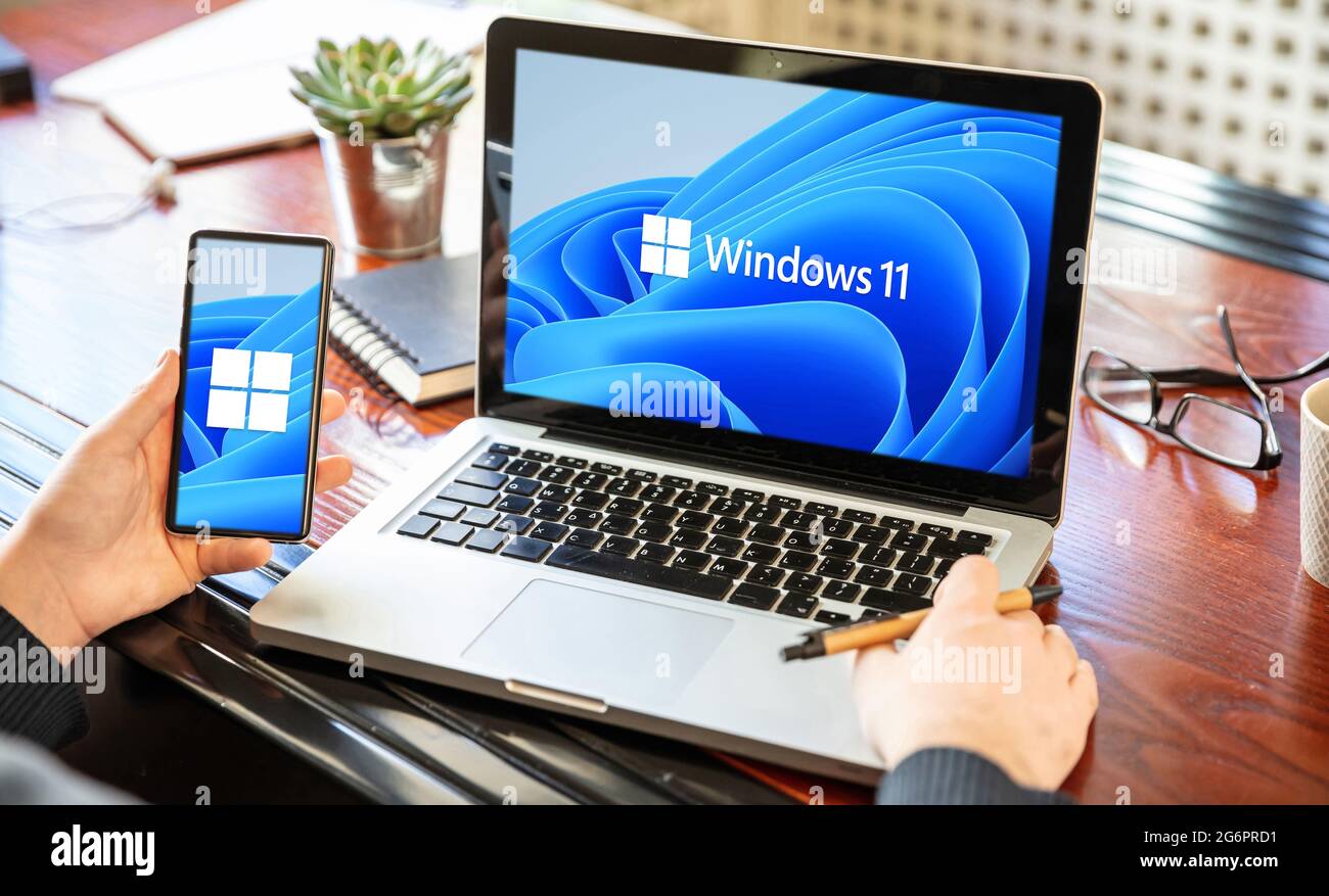 Griechenland Athen, Juli 8 2021. Mann mit einem Laptop und einem Smartphone, Windows 11 neues Microsoft-Betriebssystem auf den Bildschirmen, Business Office Schreibtisch Backgro Stockfoto