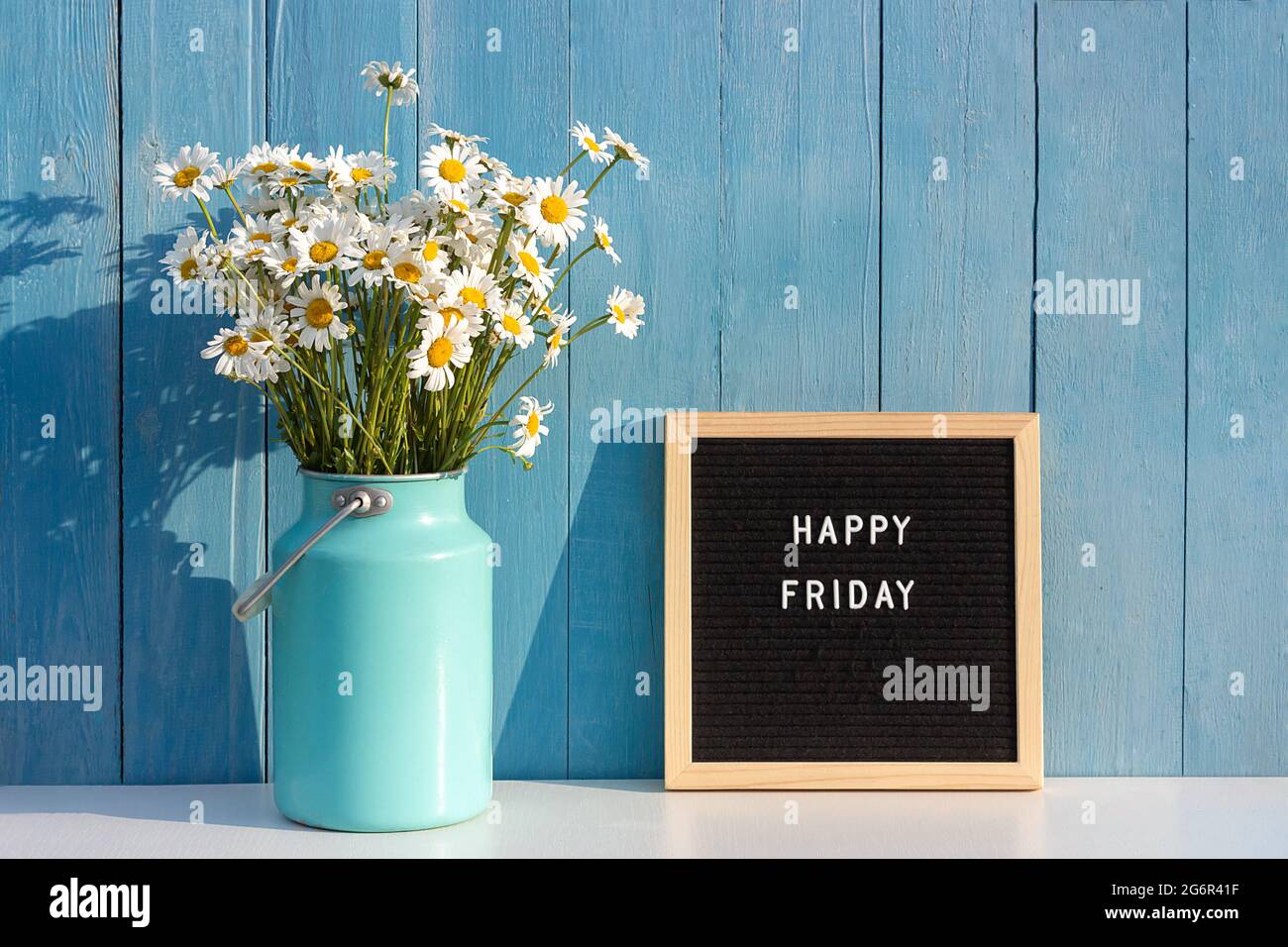 Happy Friday Worte auf schwarzem Briefbrett und Bouquet von Kamillenblumen auf dem Tisch gegen blaue Holzwand. Konzept Hallo Freitag. Stockfoto