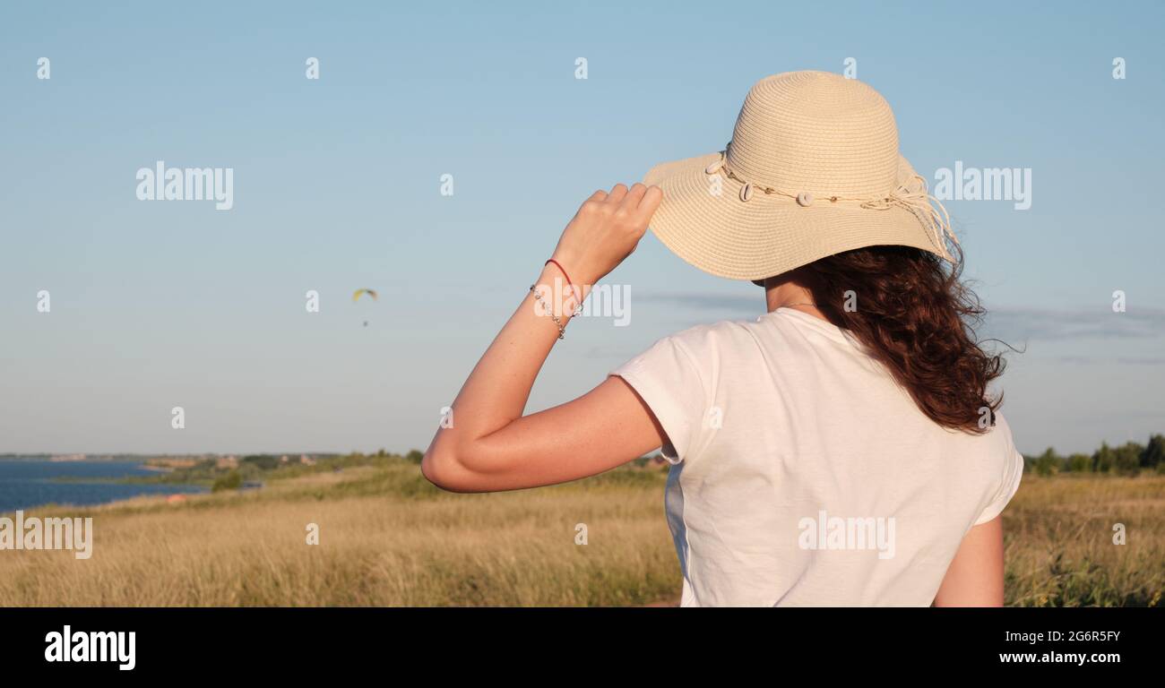 Das Mädchen steht mit dem Rücken und hält ihren Hut und schaut zum Himmel auf den Gleitschirm. Die Frau steht mit ihrem Rücken in einem Hut auf einem Hügel. Stockfoto