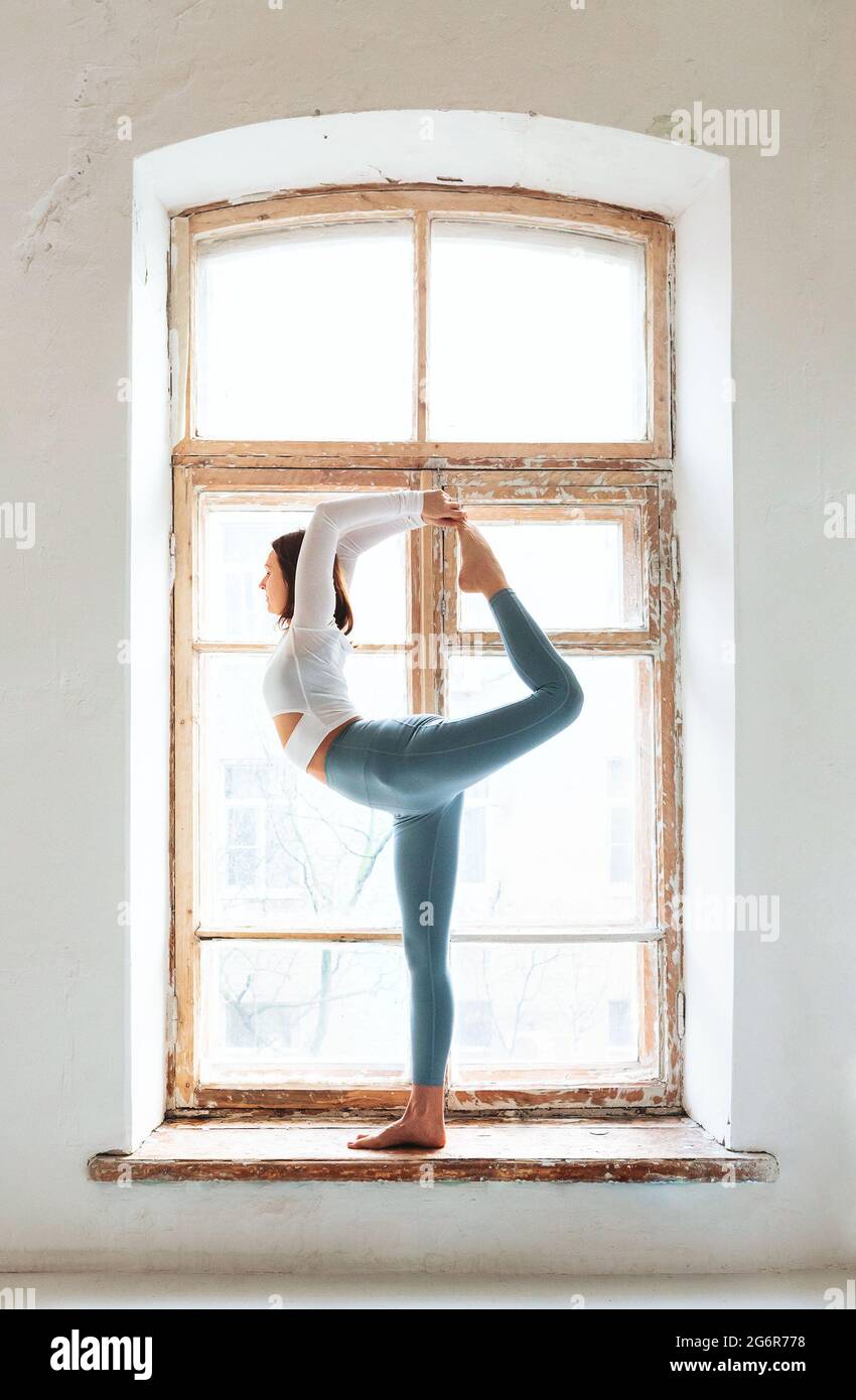 Seitenansicht einer anmutigen Frau in Sportswear, die Lord of the Dance in der Nähe eines verwitterten Fensters im hellen Raum posiert Stockfoto