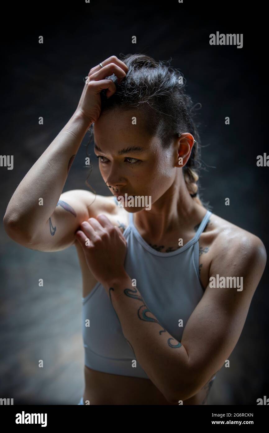 Eine junge Frau mit Tattoos in Gymnastik hält einen Arm auf ihrem Kopf, der andere wird angehoben. Sie macht eine Pause vom Training. Stockfoto