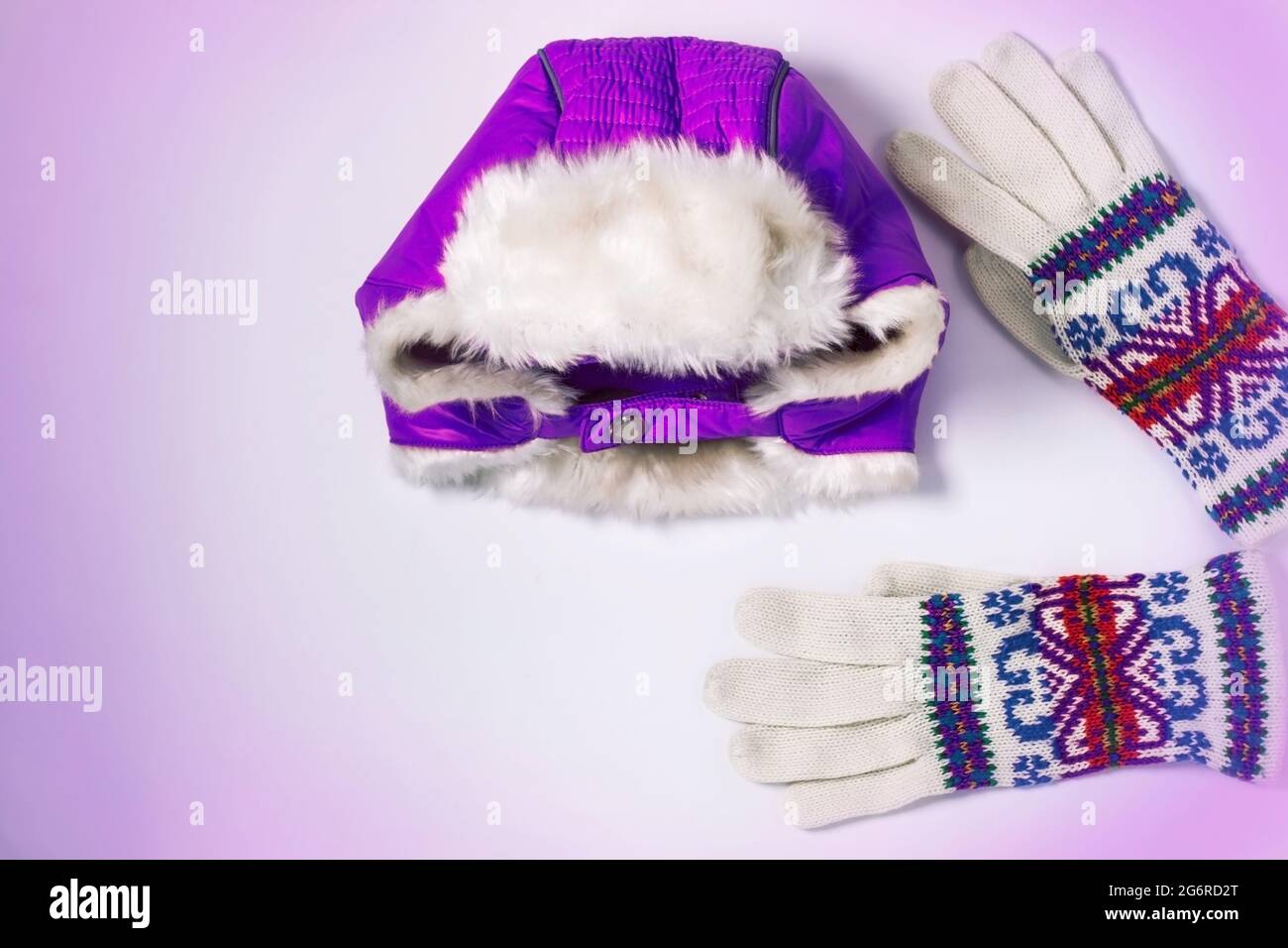 Winter-Flatlay, warmer lila Hut mit Ohrenklappen und bunten Wollgestrickten Handschuhen auf weißem violettem Hintergrund. Draufsicht, Modell für Markennamen, Kopie en Stockfoto