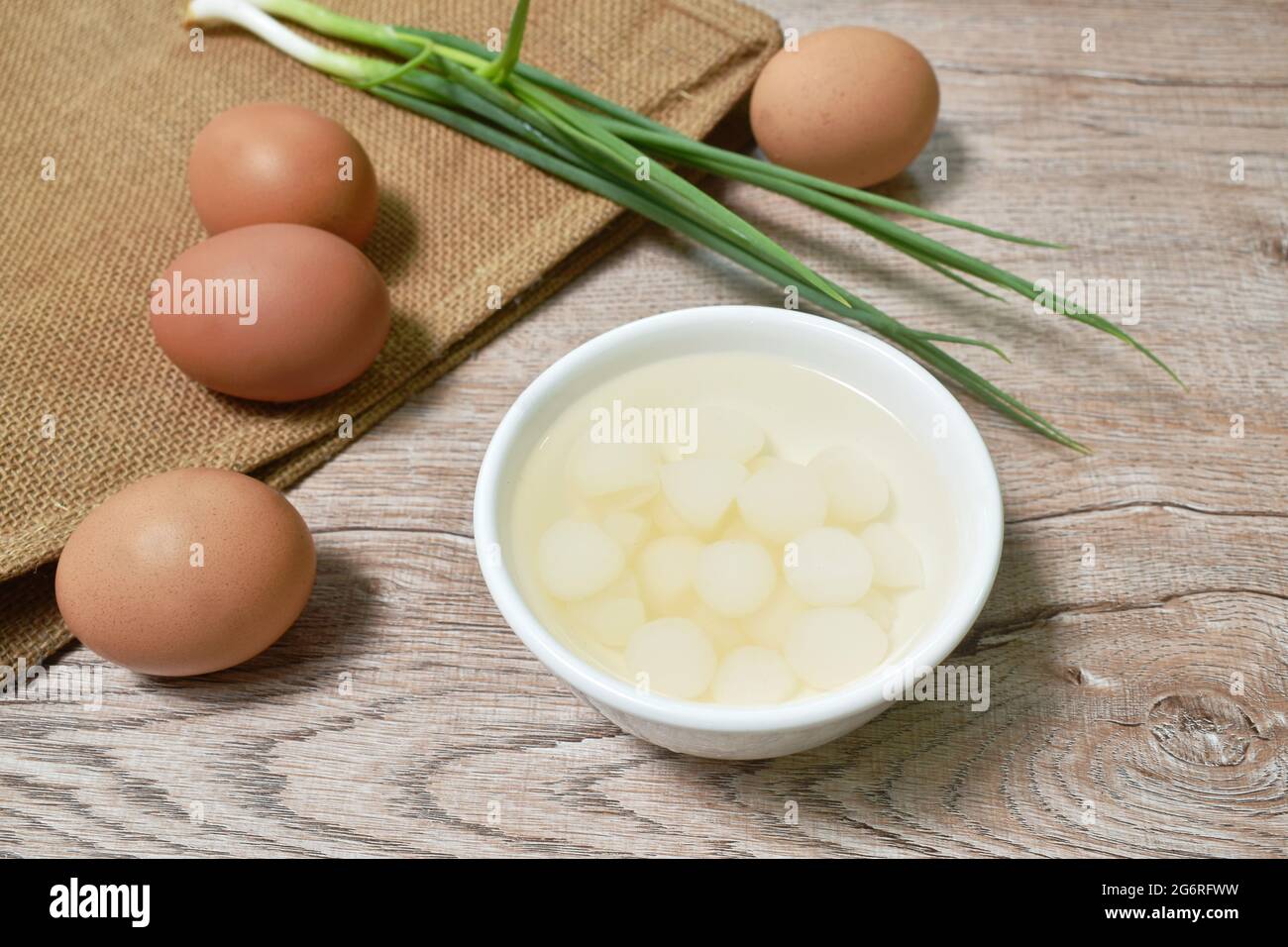 Eingelegter Knoblauch in Salzwasser auf Tasse mit rohem Ei und Frühlingszwiebeln auf dem Tisch Stockfoto
