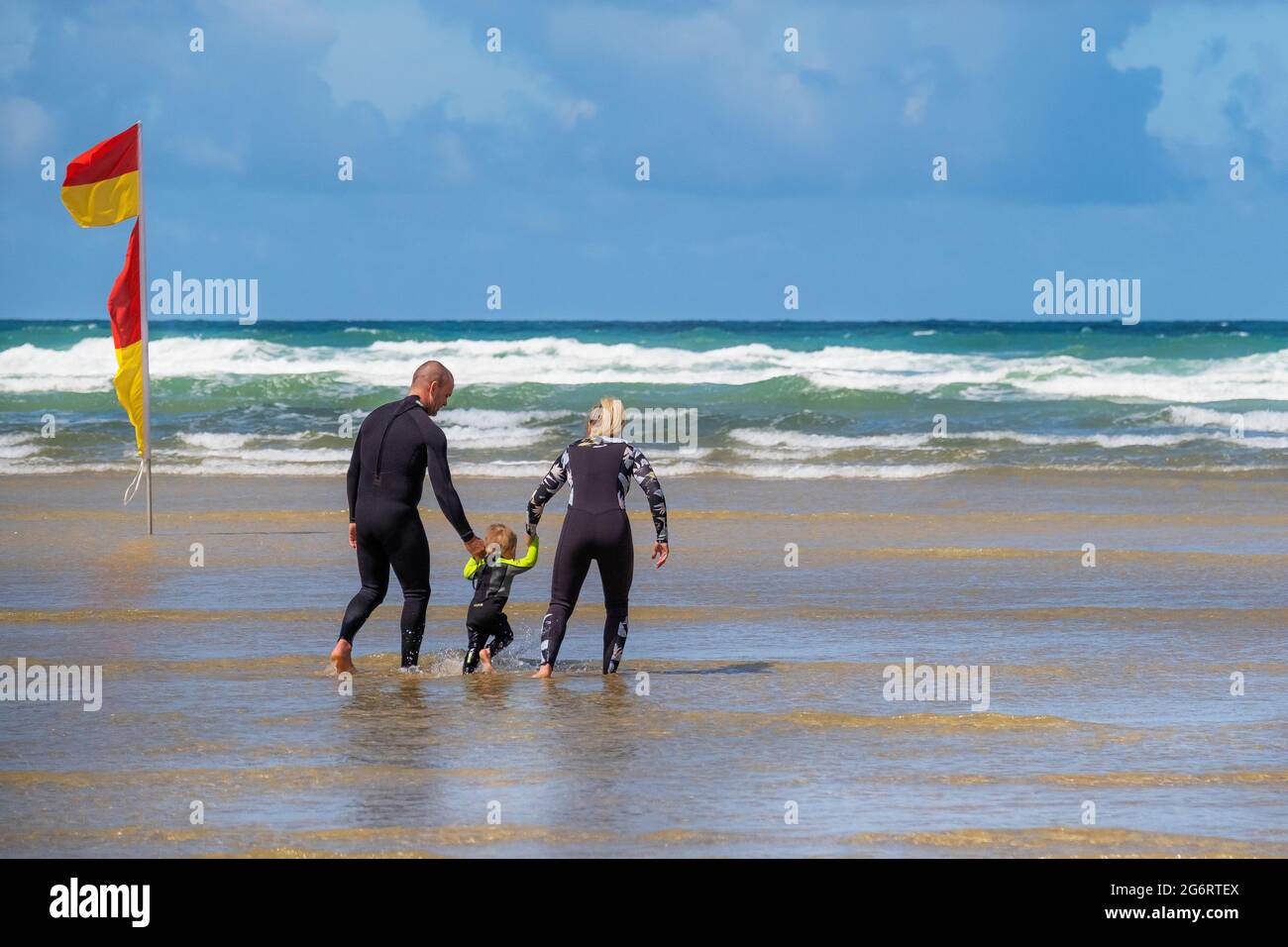 Eine Mutter und ein Vater halten die Hände mit ihrem Kleinkind, das im Meer bei Mawgan Porth in Cornwall paddelt; ein Urlaub für Familien in Cornwall. Stockfoto