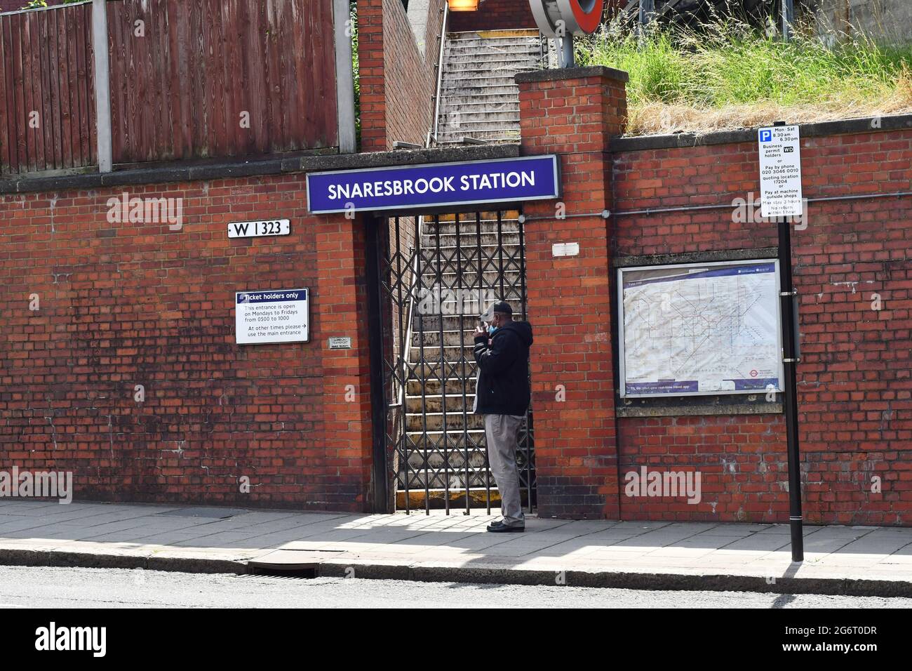 Ein Mann in seiner Maske bläst seine Nase und steht im Juli 2021 an einem geschlossenen Tor am Bahnhof Snaresbrook Stockfoto