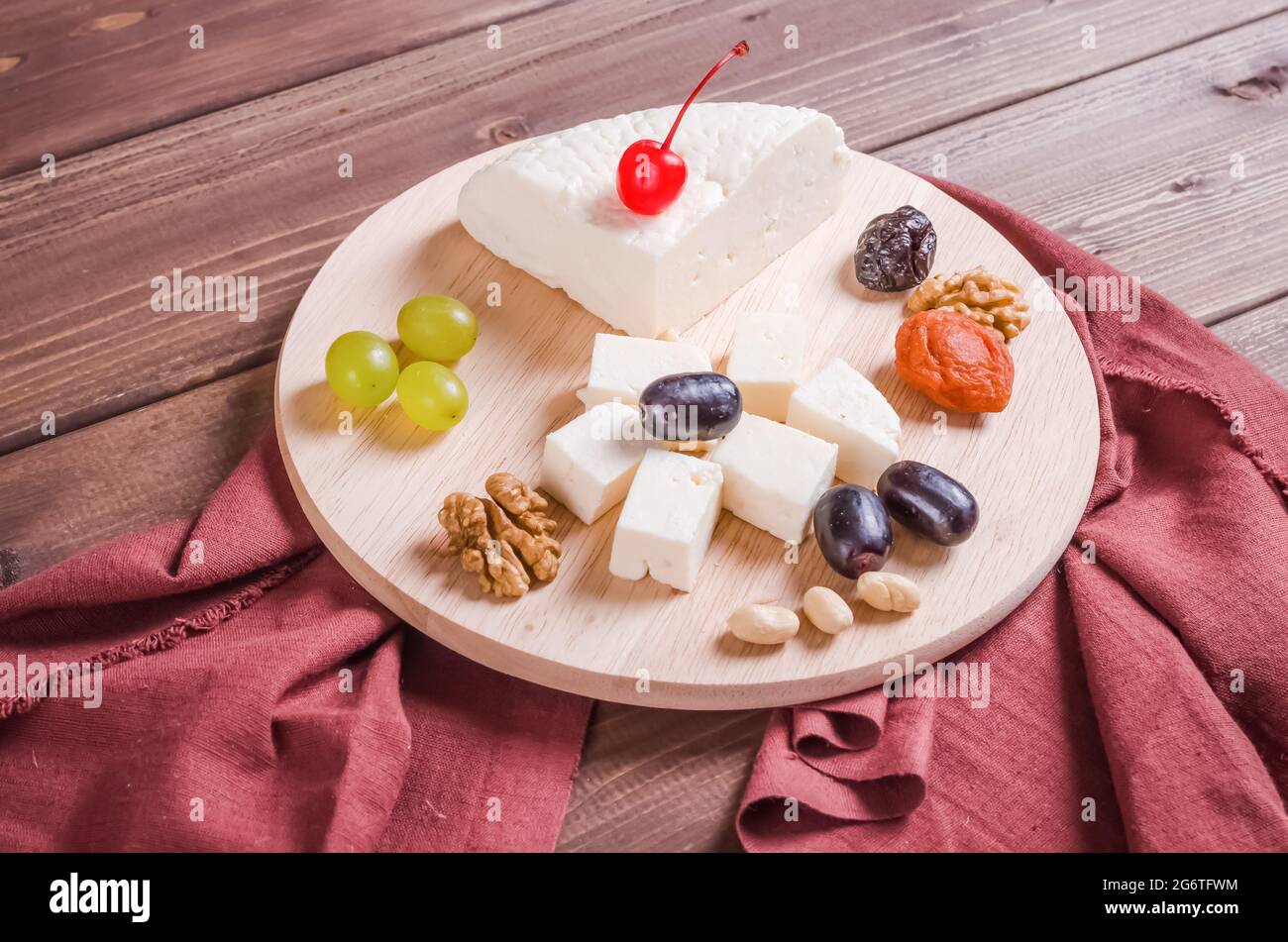 Frischer, schmackhafter, in Scheiben geschnittener adyghe-Käse, garniert mit Nüssen und Trauben auf einem runden Holzbrett auf dunklem Holzhintergrund Stockfoto