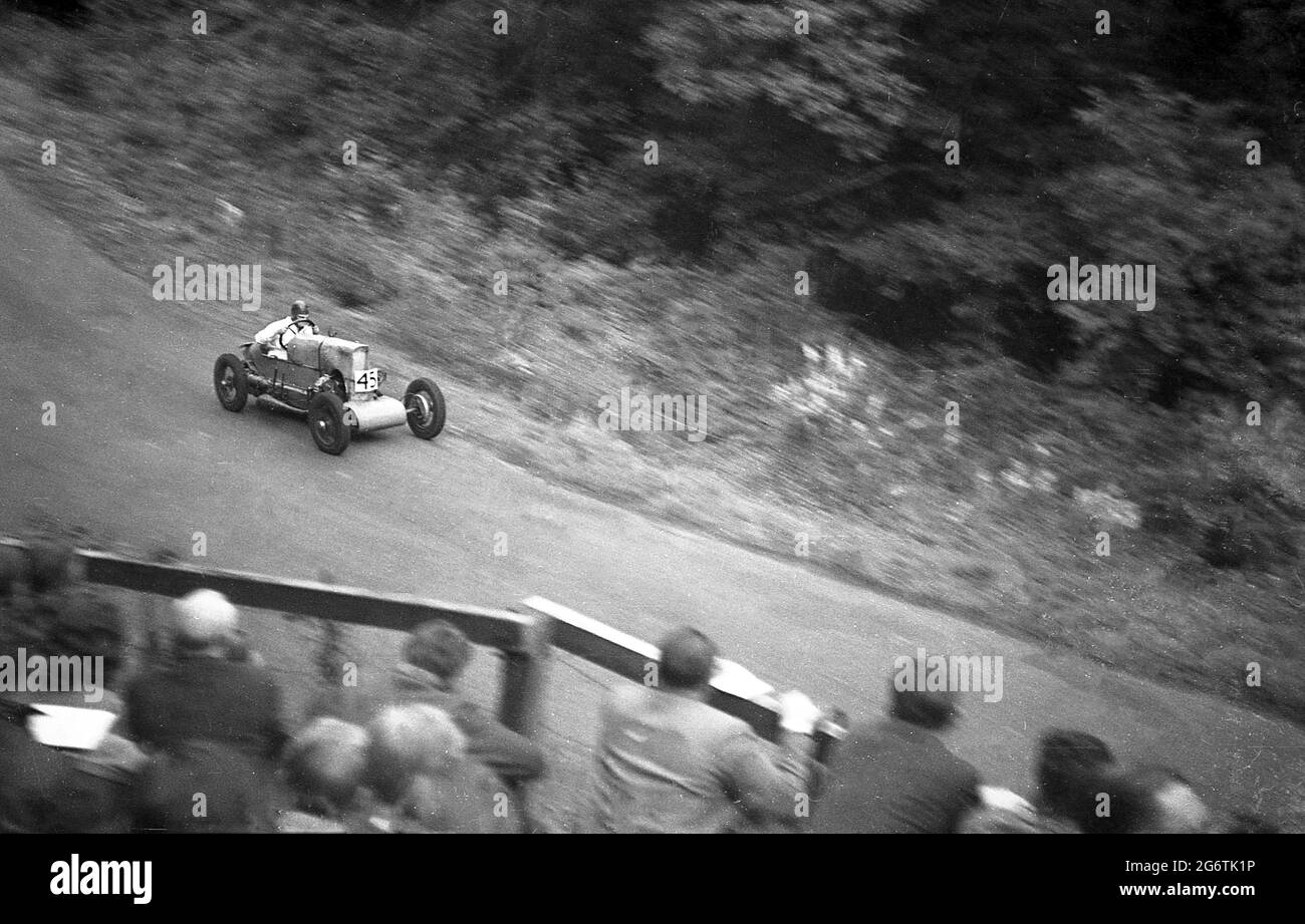 Sechziger Jahre, historische, außerhalb Motorsport-Veranstaltung, Zuschauer auf Holzbalkon beobachten von oben als ein alter Open Top-Rennwagen aus den 1920er Jahren, kommen rasenden entlang einer gepflasterten Strecke neben einem Wald, England, Großbritannien. Stockfoto