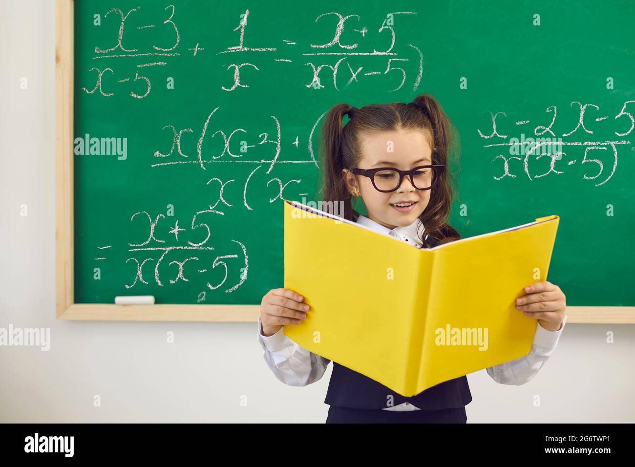 Zurück zur Schule. Intelligentes Schulmädchen in einer Brille mit einem Buch, das auf dem Hintergrund einer grünen Tafel in einer Schulklasse steht Stockfoto
