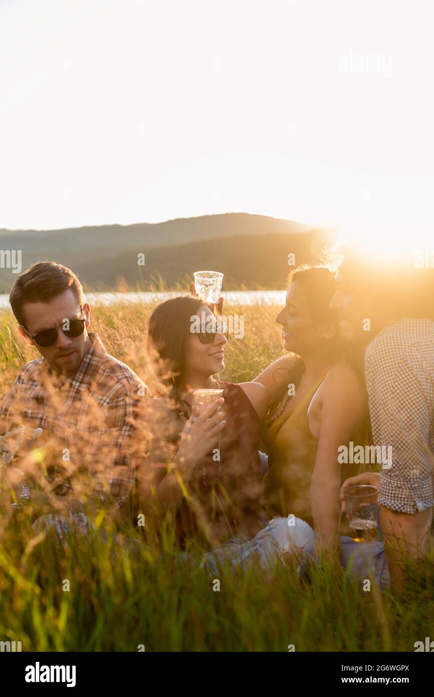 Freunde genießen die Abendstimmung Sommertages sitzen im hohen Grass mit Getränken und Gitarrenmusik Stockfoto