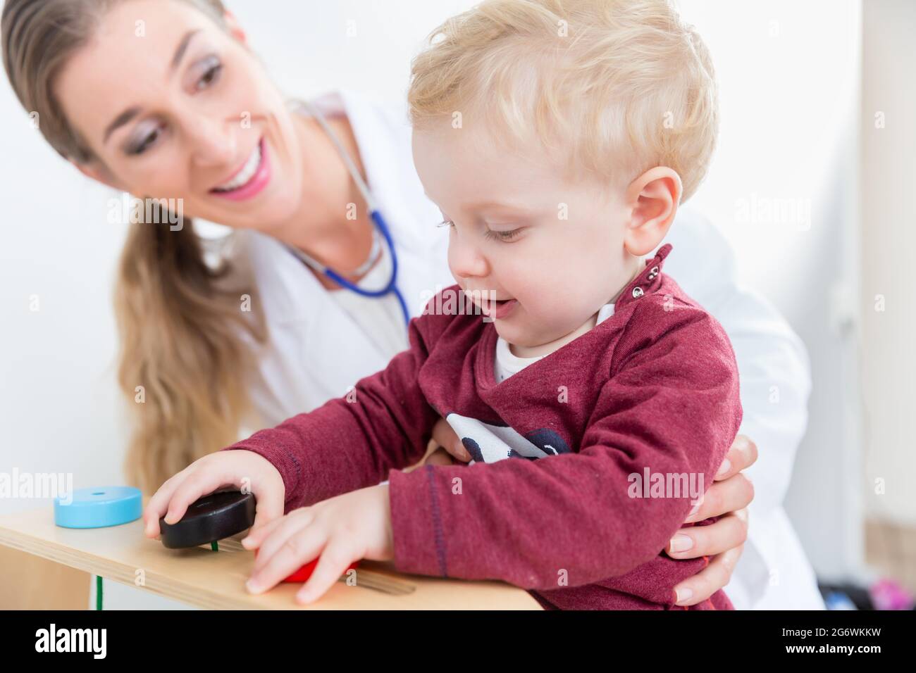 Netter aktiver Junge, der mit Holzspielzeug in den Armen einer erfahrenen Kinderärztin während der routinemäßigen körperlichen Untersuchung in einer modernen Medica spielt Stockfoto