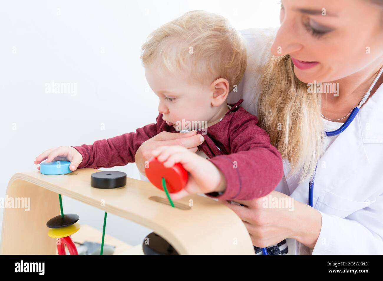 Netter aktiver Junge, der mit Holzspielzeug in den Armen einer erfahrenen Kinderärztin während der routinemäßigen körperlichen Untersuchung in einer modernen Medica spielt Stockfoto