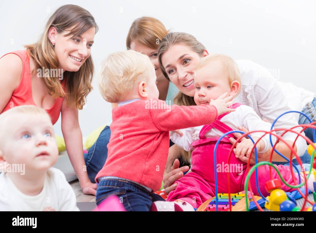 Eine Gruppe von glücklichen jungen Müttern, die ihre süßen und gesunden Babys beobachten, während sie mit bunten Spielsachen in einer modernen Kindertagesstätte spielen Stockfoto