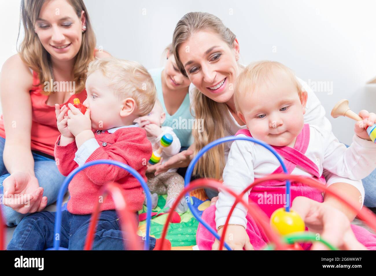 Eine Gruppe von glücklichen jungen Müttern, die ihre süßen und gesunden Babys beobachten, während sie mit bunten Spielsachen in einer modernen Kindertagesstätte spielen Stockfoto