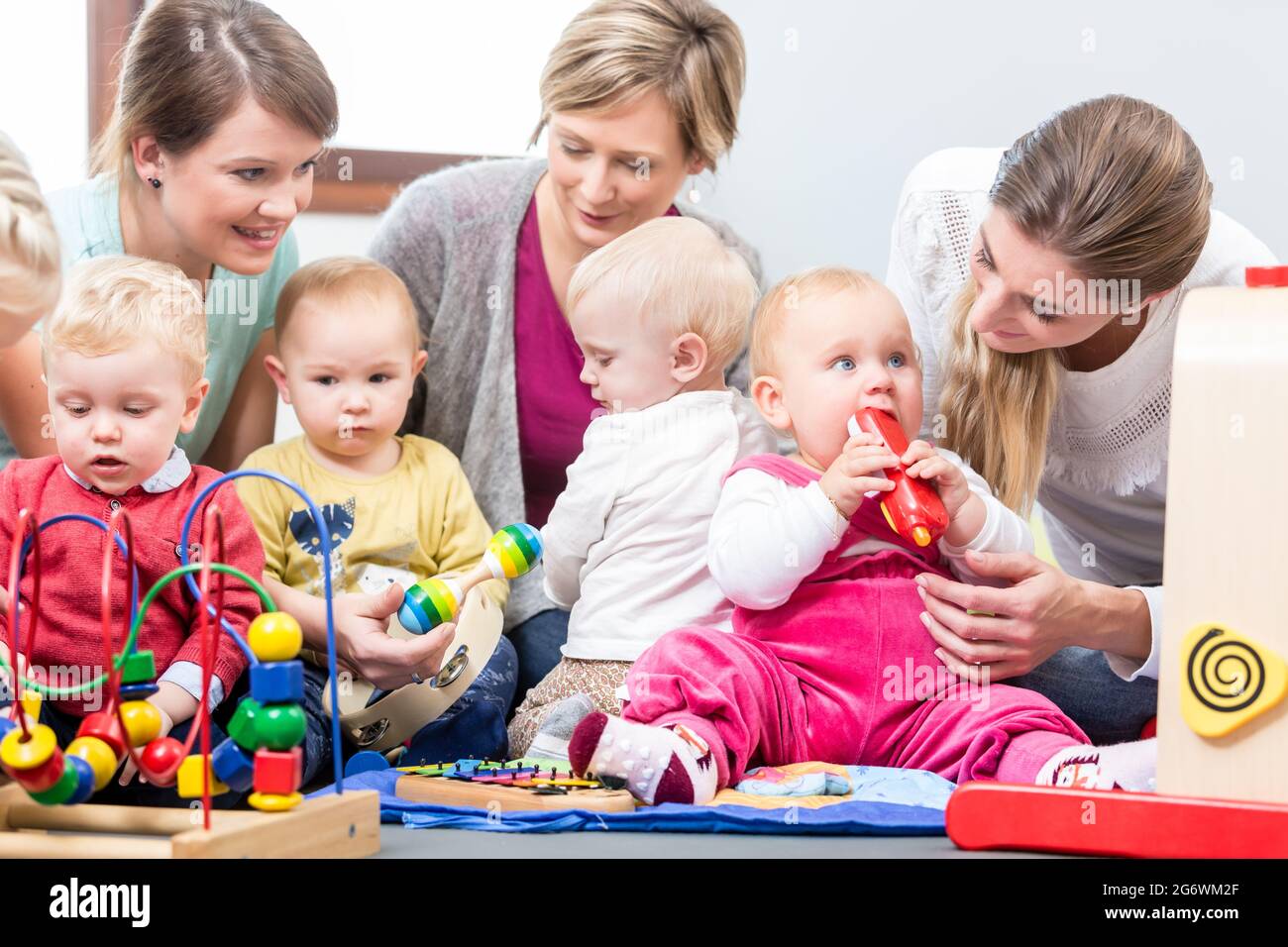 Drei engagierte und glückliche junge Mütter sitzen zusammen auf dem Boden, während sie ihren Babys beim Spielen mit sicheren bunten Spielsachen in einer modernen pla zusehen Stockfoto