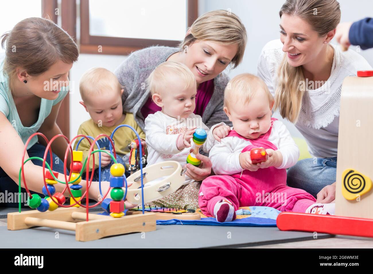 Drei engagierte und glückliche junge Mütter sitzen zusammen auf dem Boden, während sie ihren Babys beim Spielen mit sicheren bunten Spielsachen in einer modernen pla zusehen Stockfoto