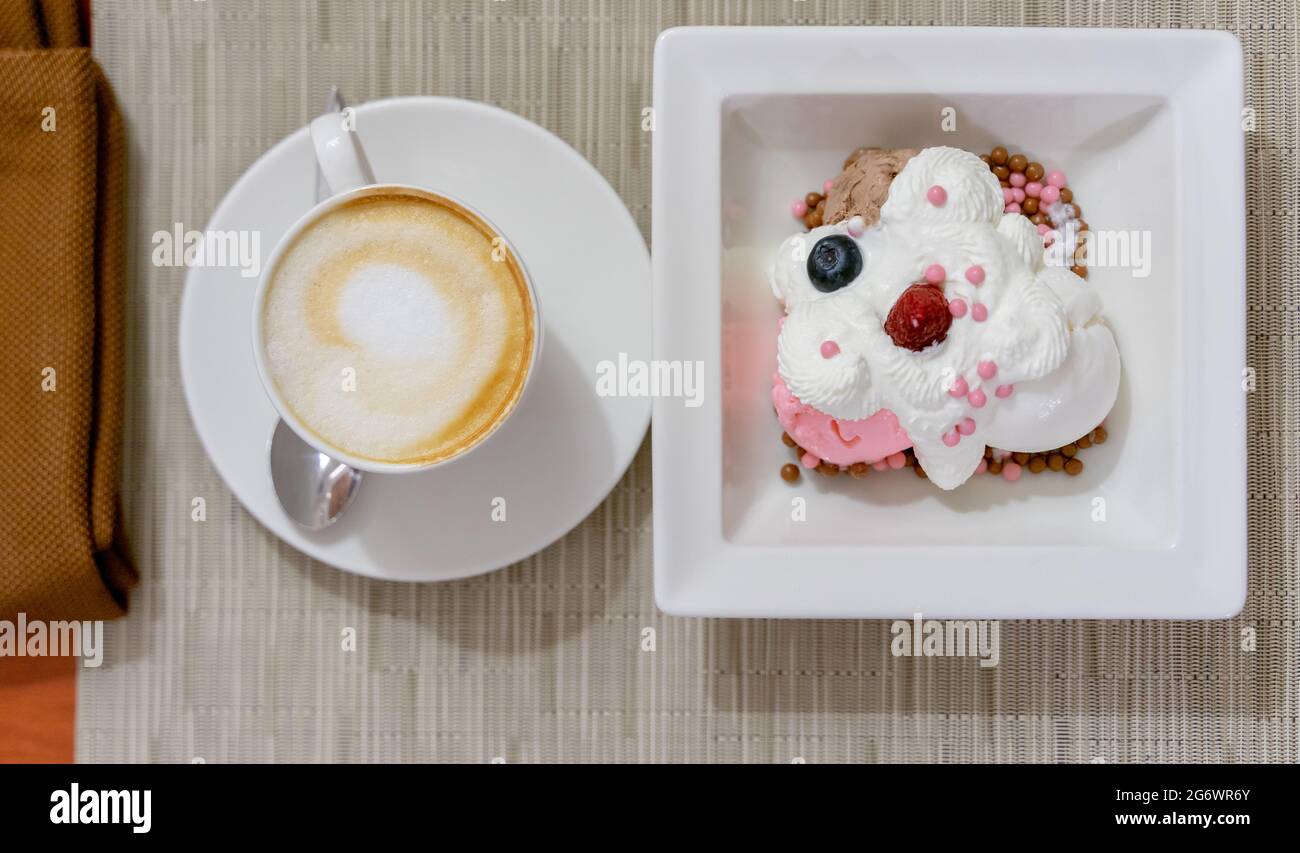 Blick von oben auf Cappuccino in einer weißen Tasse und Vanille- und Erdbeereis mit Schlagsahne und Beeren auf einem weißen Dessertgericht. Stockfoto