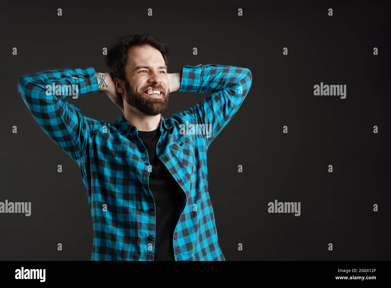 Bärtig lächelnder Mann im Hemd posiert mit Händen hinter seinem Hals auf schwarzem Hintergrund isoliert geklammert Stockfoto