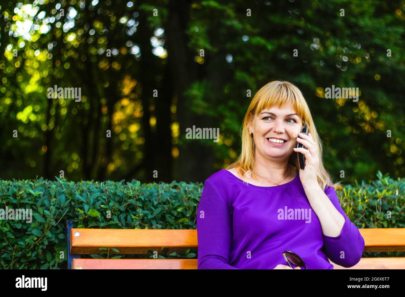 Unschärfe kaukasisch blonde lächelnde Frau sprechen, sprechen auf dem Telefon draußen, draußen. 40-jährige Frau in lila Bluse im Park auf der Bank. Erwachsene fe Stockfoto