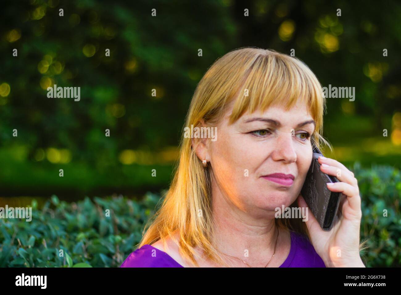 Unschärfe-Nahaufnahme kaukasisch blonde Frau sprechen, sprechen auf dem Telefon draußen, im Freien. 40-jährige Frau in lila Bluse im Park. Erwachsene weibliche Erbsenw Stockfoto