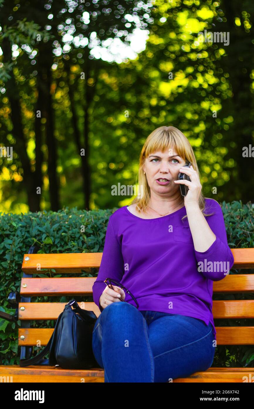 Unschärfe wütend kaukasisch blonde Frau reden, sprechen am Telefon draußen, im Freien. 40-jährige Frau in lila Bluse im Park. Erwachsene Person mit Stockfoto
