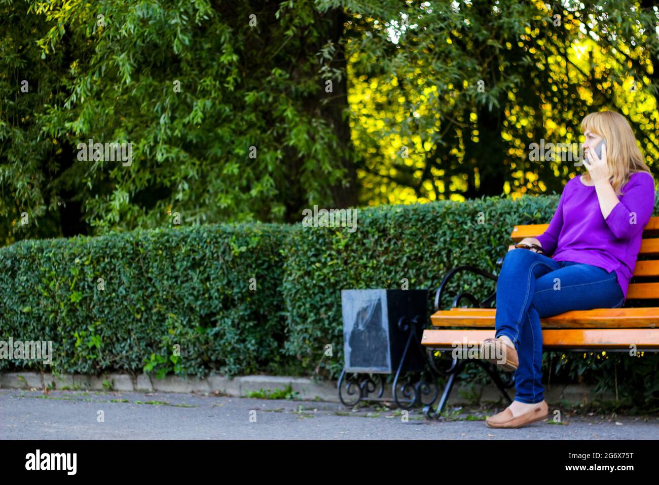 Unschärfe kaukasisch blonde Frau reden, sprechen am Telefon draußen, im Freien. 40-jährige Frau in lila Bluse im Park. Erwachsene weibliche Person, die ein Mobiltelefon verwendet Stockfoto