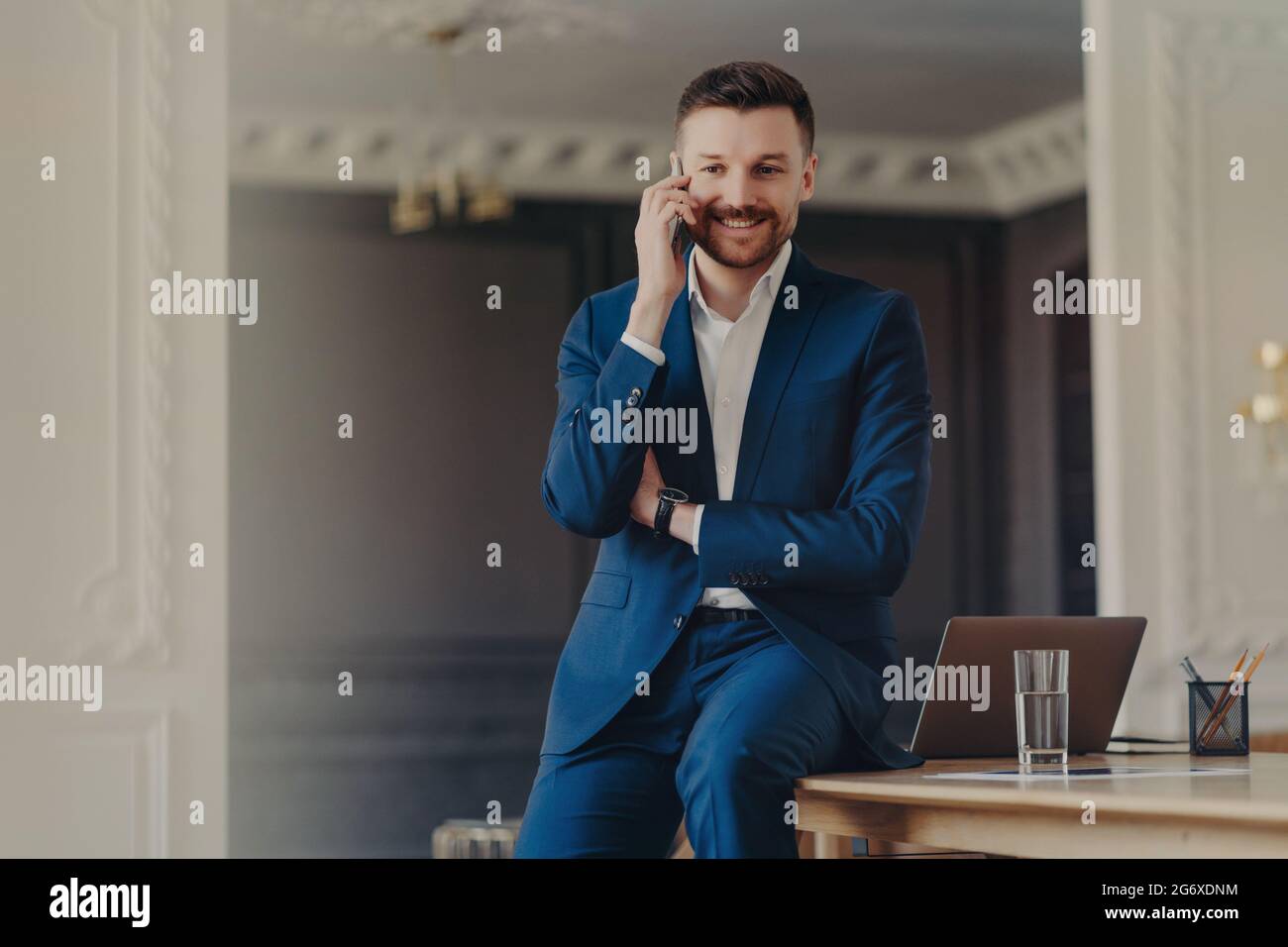 Das Foto eines gut aussehenden bärtigen Mannes mit Telefongesprächen macht den Anruf auf dem Mobiltelefon mit guten Nachrichten auf dem Desktop mit einem modernen Laptop-Computer zufrieden Stockfoto