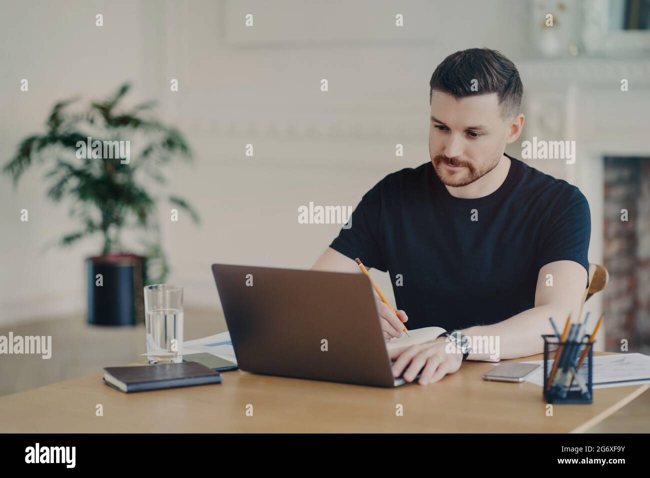 Seriös bärtiger Mann schreibt informative Notizen, während er sich das Webinar auf einem Laptop-Computer ansieht, der in lässigen T-Shirt-Posen am Arbeitsplatz gekleidet arbeitet Stockfoto