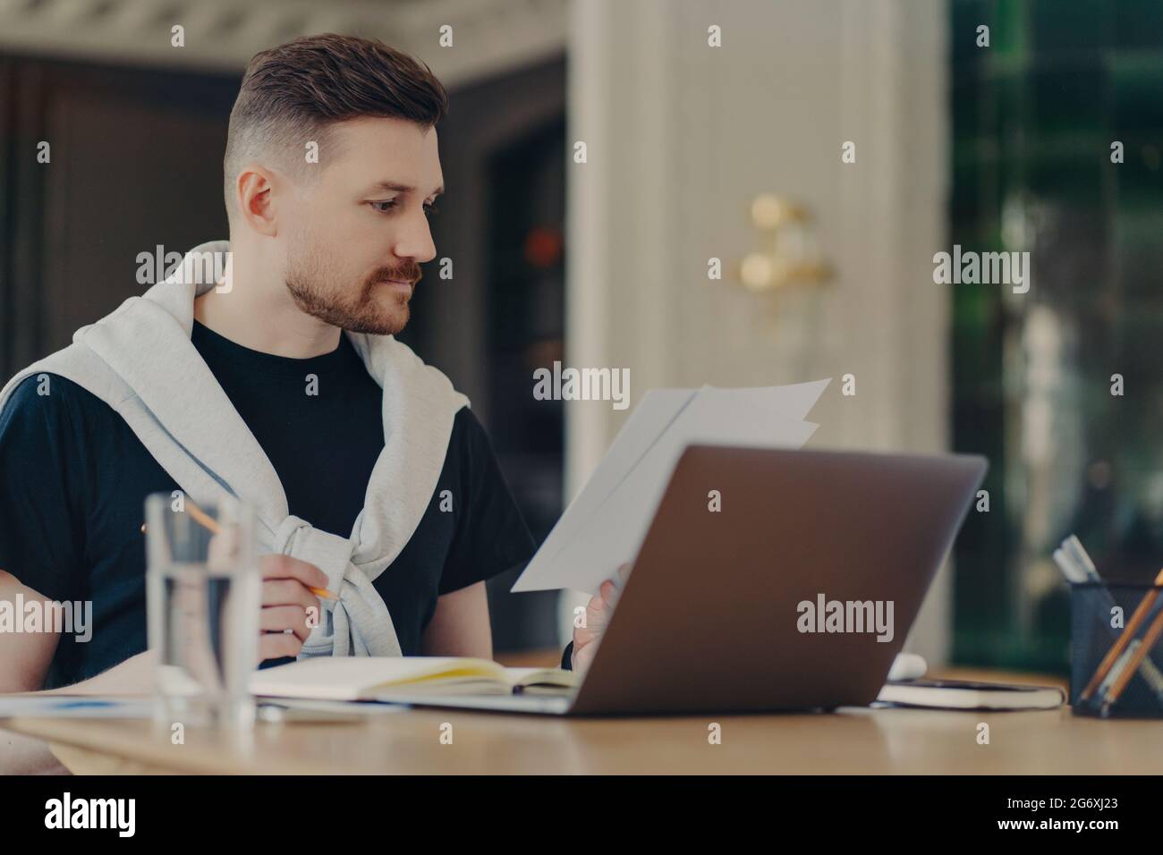 Smart Handsome Mann prüft Informationen auf Papieren funktioniert Remote sitzt zu Hause Büro verwendet modernen Laptop-Computer schreibt in Notebook gekleidet Stockfoto