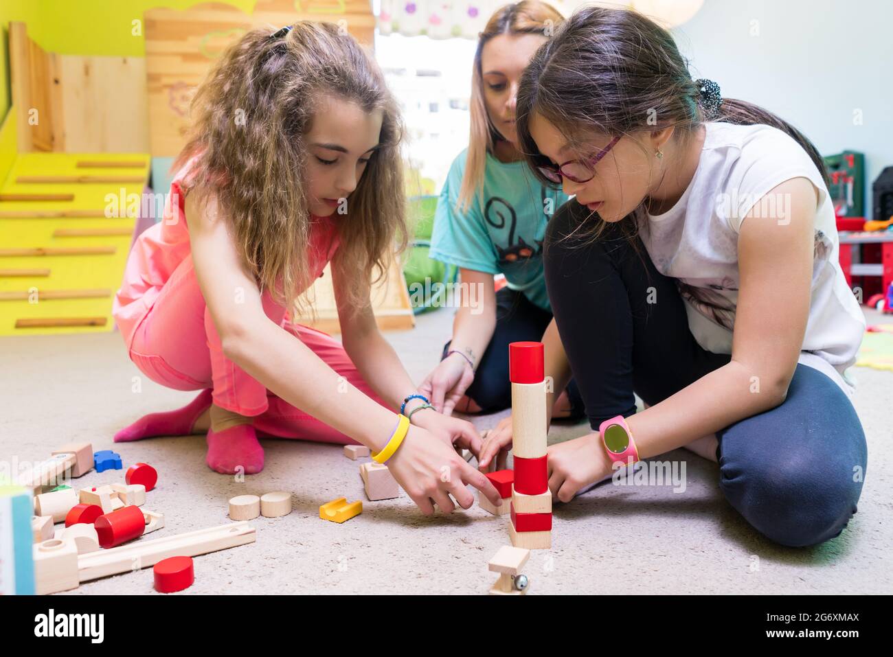 Zwei Pre-school Mädchen zusammen spielen mit Holzspielzeug Bausteine auf dem Boden während der Spielzeit betreut durch eine sorgfältige junge Kindergärtnerin Stockfoto