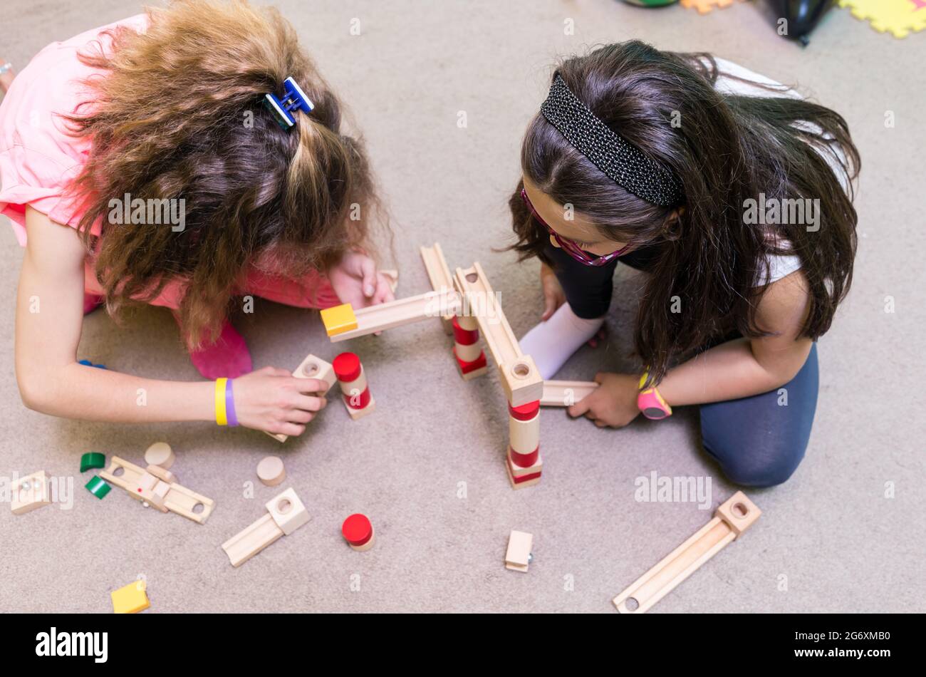 Blick aus der Perspektive von zwei Mädchen aus der Vorschule, die sich Holzspielzeugblöcke teilen, während sie im Kindergarten eine herausfordernde Struktur auf dem Boden aufbauen Stockfoto