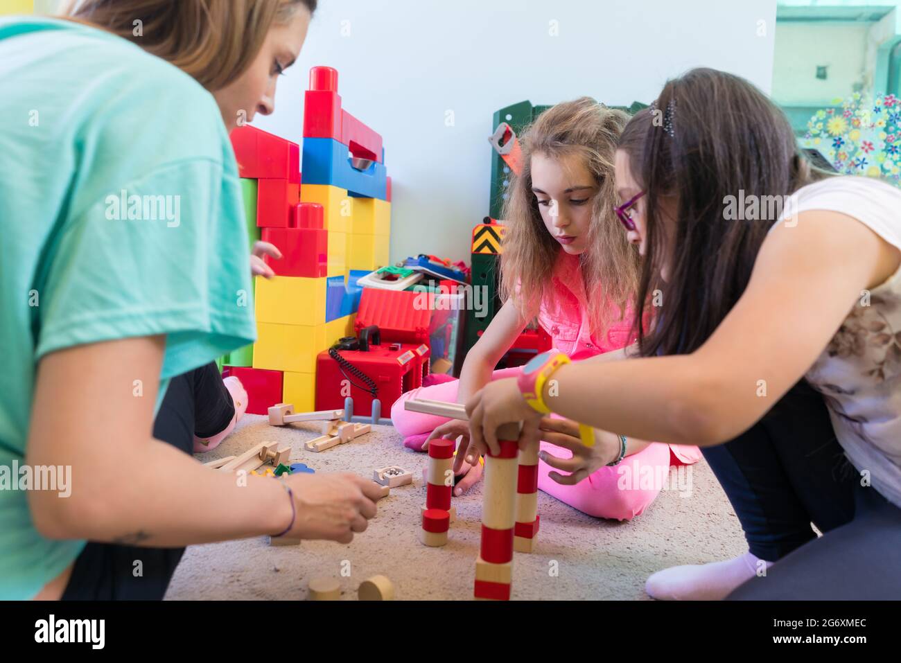 Vorschulmädchen spielen während der Spielzeit mit hölzernen Spielzeugblöcken auf dem Boden, die von einer vorsichtigen jungen Kindergärtnerin betreut werden Stockfoto