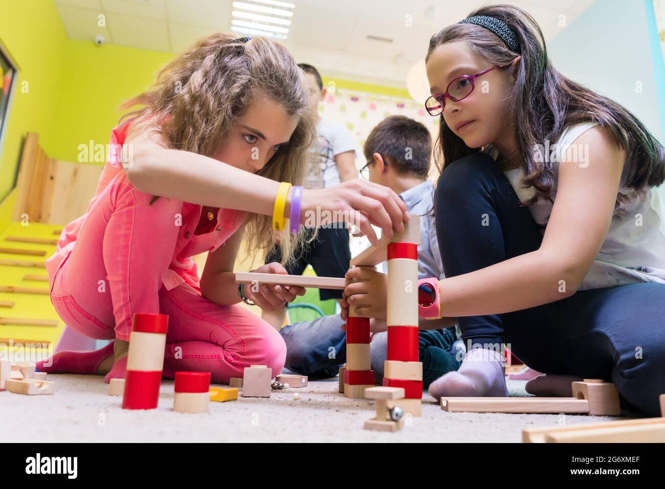 Zwei Pre-school Mädchen zusammen spielen mit Holzspielzeug Bausteine auf dem Boden während der Spielzeit betreut durch eine sorgfältige junge Kindergärtnerin Stockfoto