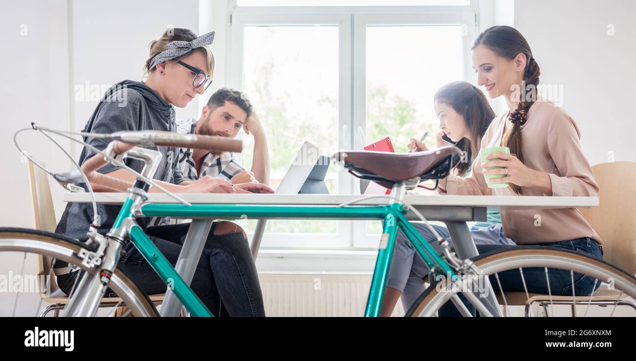 Vier kompetente Freiberufler und unabhängige Auftragnehmer arbeiten an Tablets und Laptops, die an einem gemeinsam genutzten Schreibtisch hinter einem Pendlerbi mit dem Internet verbunden sind Stockfoto