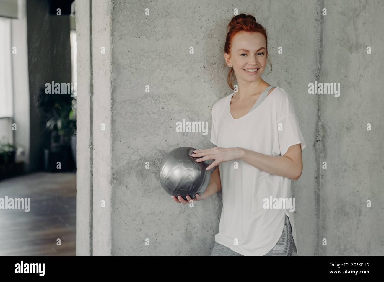 Rothaarige positive Frau, die vor der Pilates-Klasse mit einem Fitball an der grauen Wand steht Stockfoto