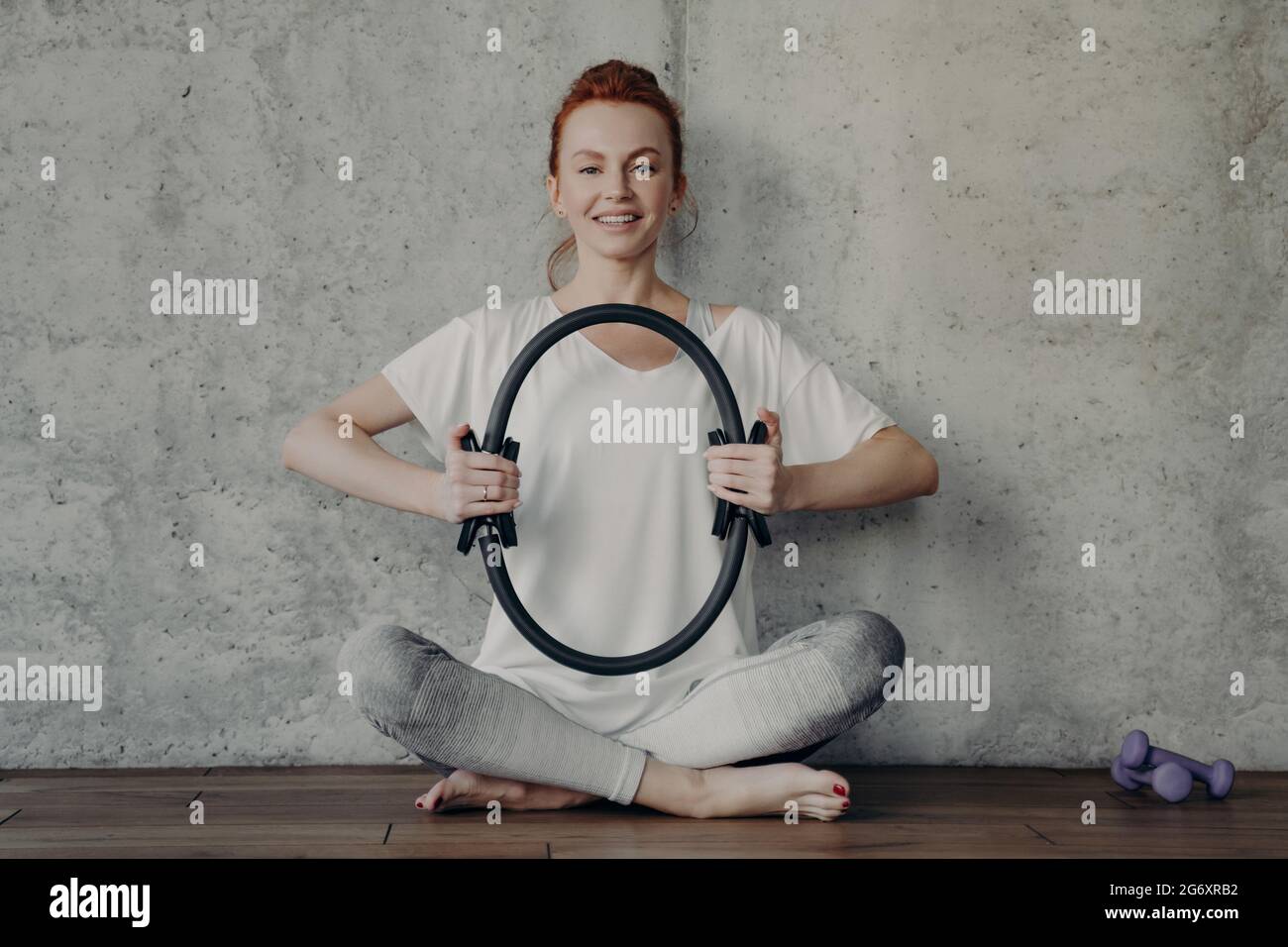 Fröhliche Fitness Frau in aktiver Kleidung sitzt in Lotusposition und trainiert mit Pilates Ring Stockfoto