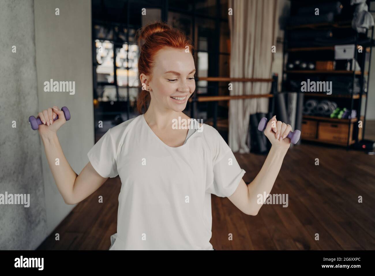 Lächelnde, sportive Rothaarige Frau trainiert mit Kurzhanteln im Fitnessstudio Stockfoto