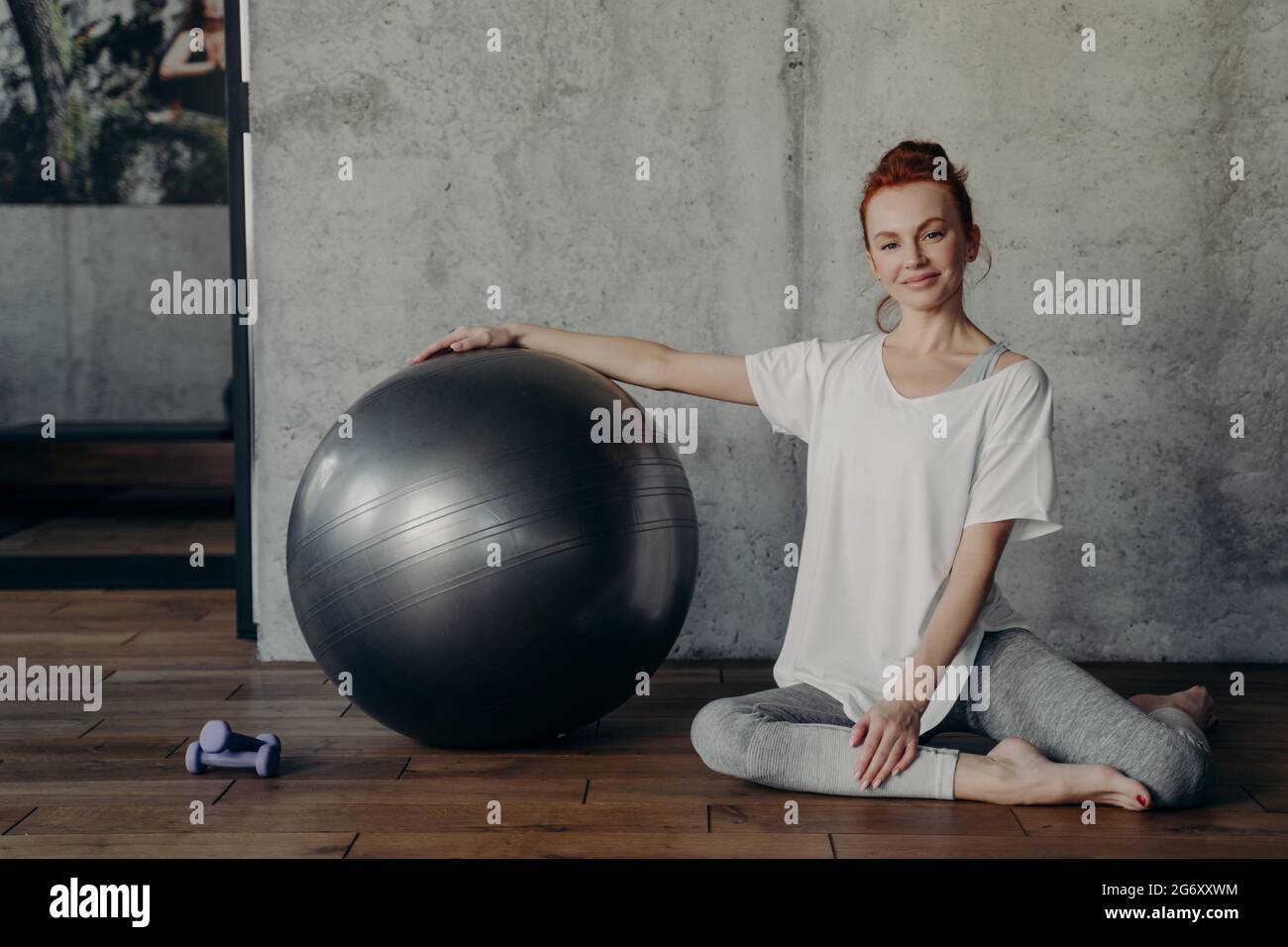 Glückliche junge Fitness-Frau, die sich nach dem Pilates-Training entspannt, während sie mit einem großen Gymnastikball auf dem Boden sitzt Stockfoto