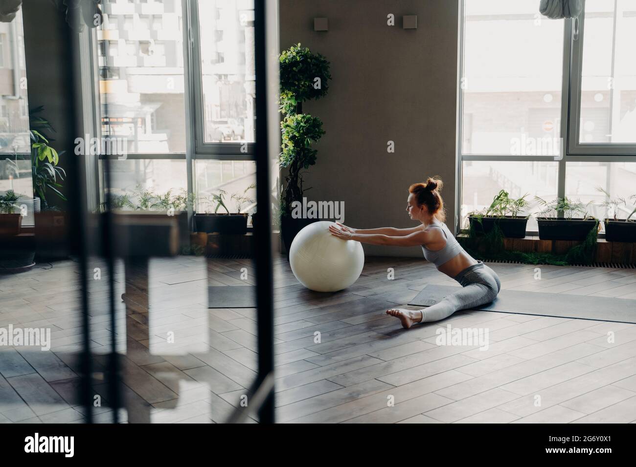 Sportliche Frau, die die Beine in geteilter Position streckt und mit einem großen Gymnastikball trainiert Stockfoto