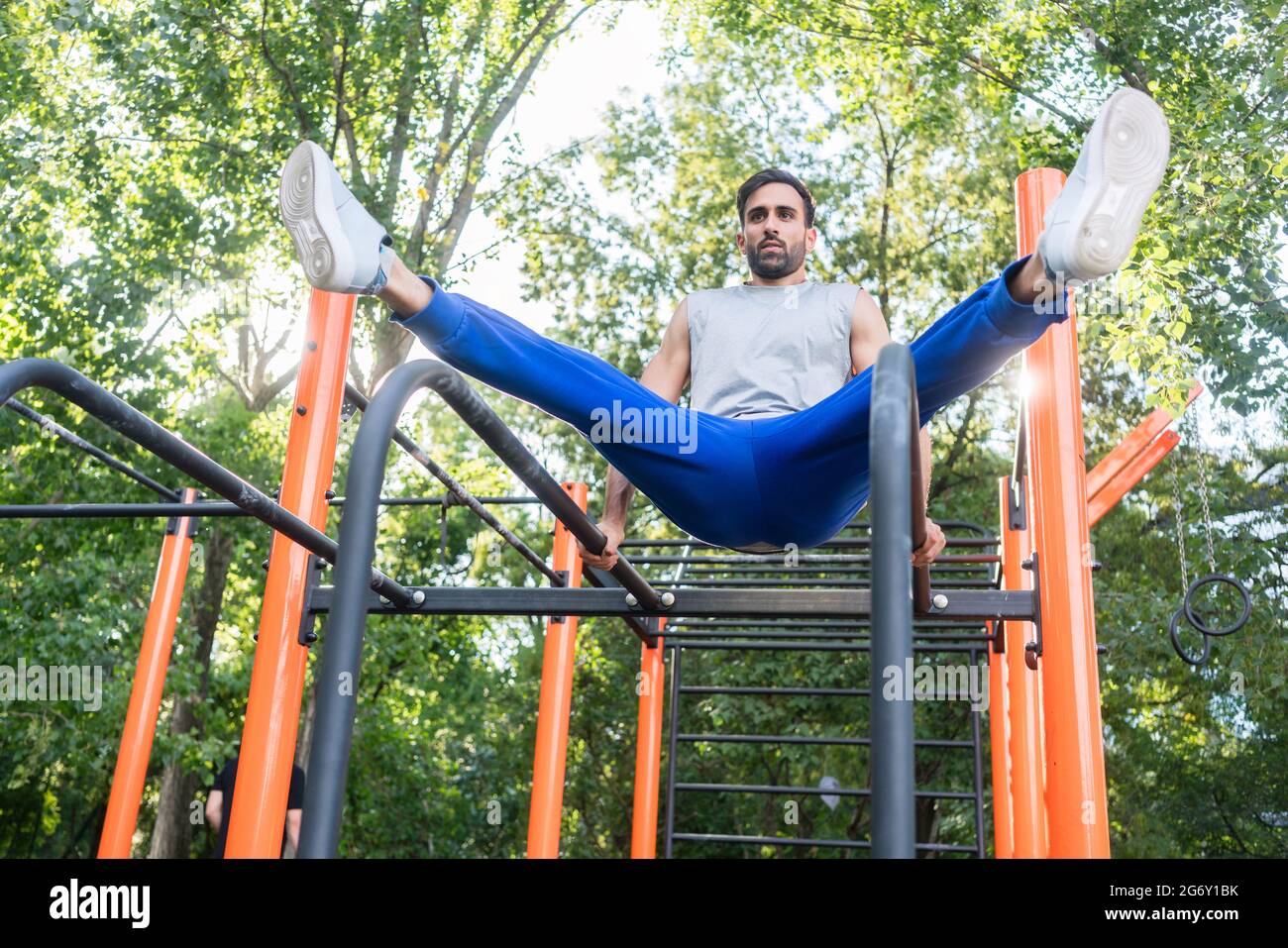 Blick aus dem unteren Winkel auf einen hübschen, athletischen jungen Mann, der in einem Outdoor-Fitnesspark mit modernen Geräten auf parallelen Gittern das vertikale Bein hebt Stockfoto